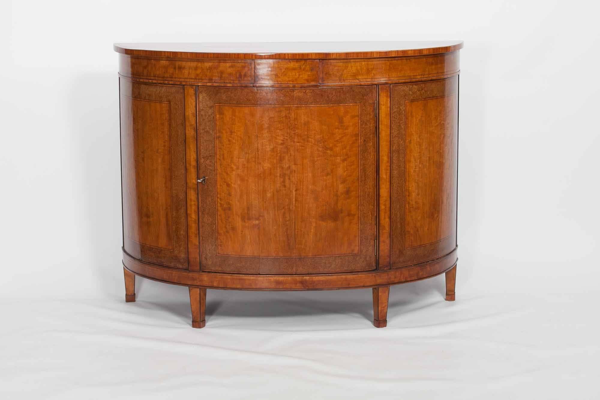 Early 19th century Regency demilune satinwood commode. The top and bow-fronted doors cross-banded with burr walnut and inlaid with fruitwood. Containing one interior shelf and terminating on four tapering legs, circa 1820.