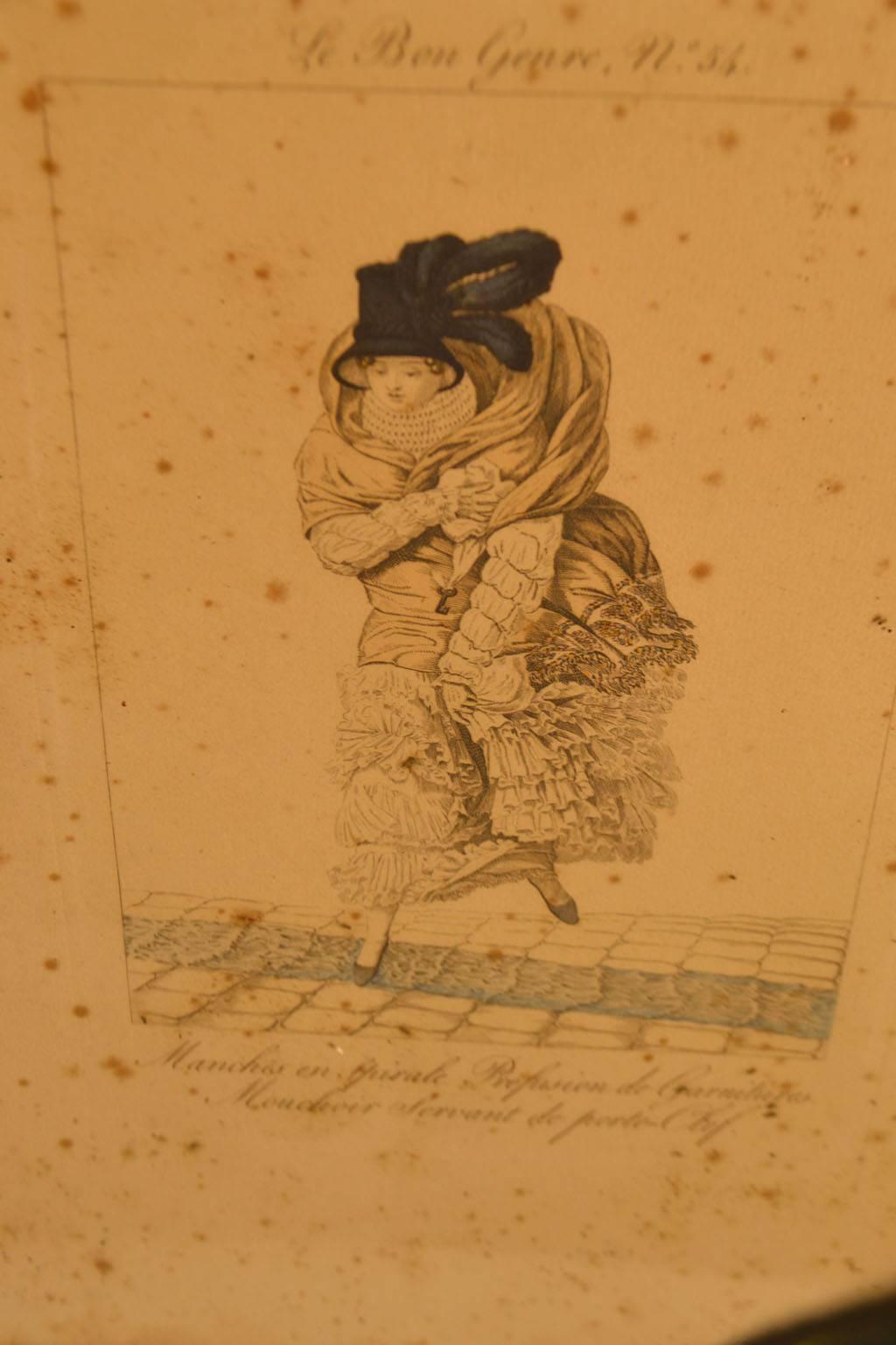 18th century framed fashion engraving of a woman in a black plume feather hat stepping off a cobblestone street in a blustery wind bundled up to keep warm. A good amount of pitting to this engraving is noticeable.