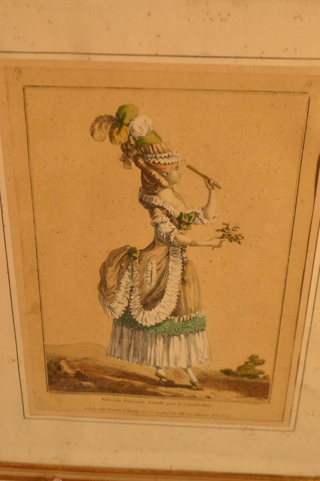gerahmter Modestich aus dem 18. Jahrhundert, der eine Frau zeigt, die zur Unterhaltung oder zur Bewirtung gekleidet ist, mit einem Federhut, einer schicken Bustle mit grüner Borte und einem Blumenstrauß in der Hand.