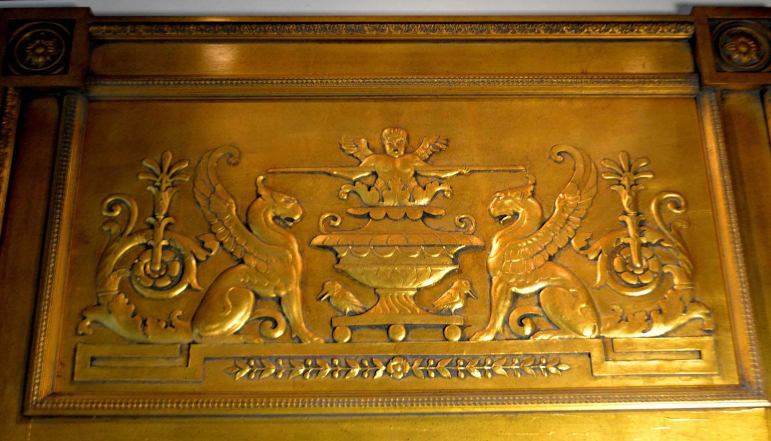 Empire-style giltwood looking glass, the frame with projecting floral block-carved corners surrounding a carved frieze panel depicting griffins flanking an urn issuing a putti figure, with the beveled rectangular plate below.