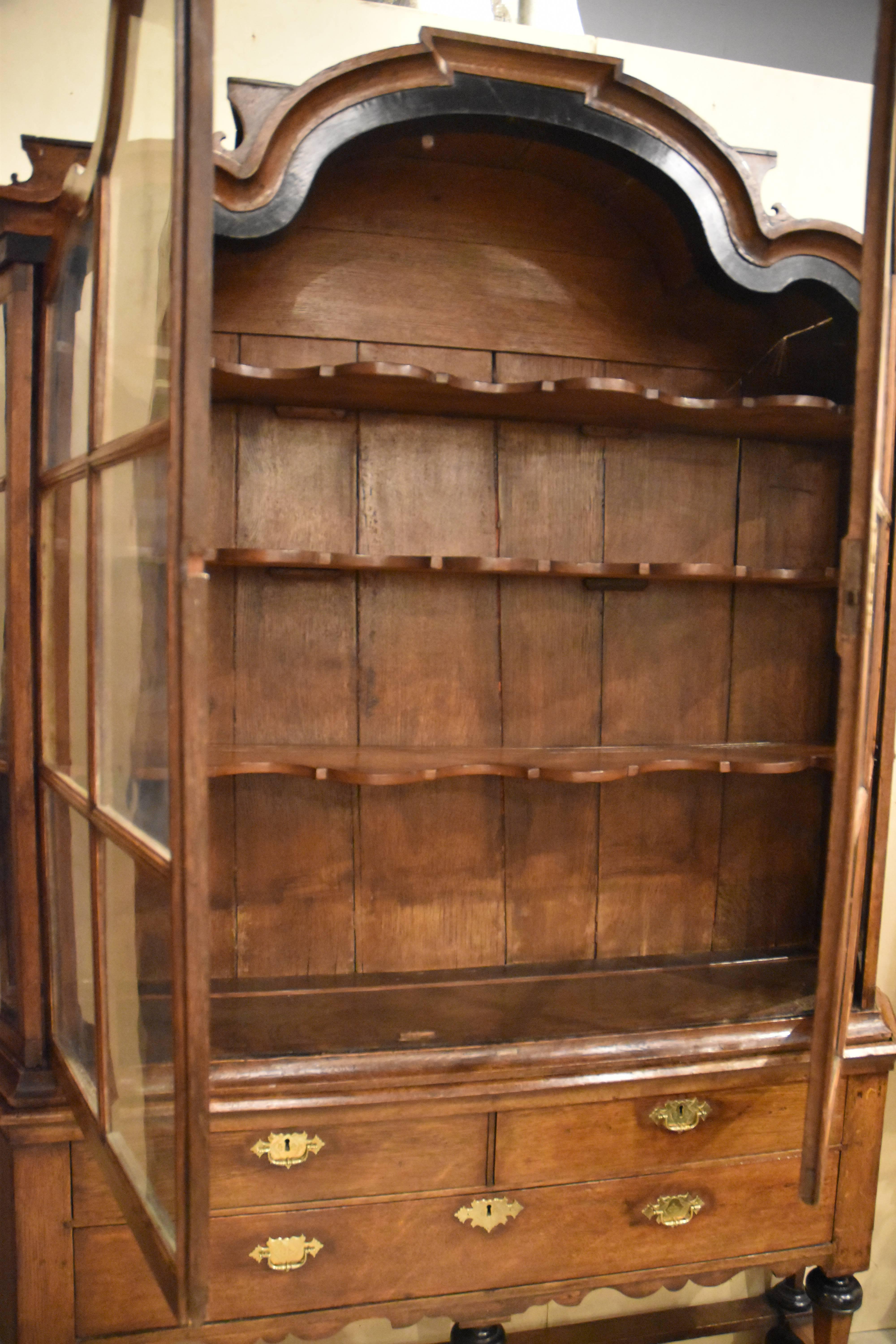 Dutch oak vitrine topped by porcelain shelves atop a serpentine shaped crest with decorative ebonized ribbon banding above conforming door frames. Double doors each have six panes, most with original glazing. The canted sides are glazed, each with