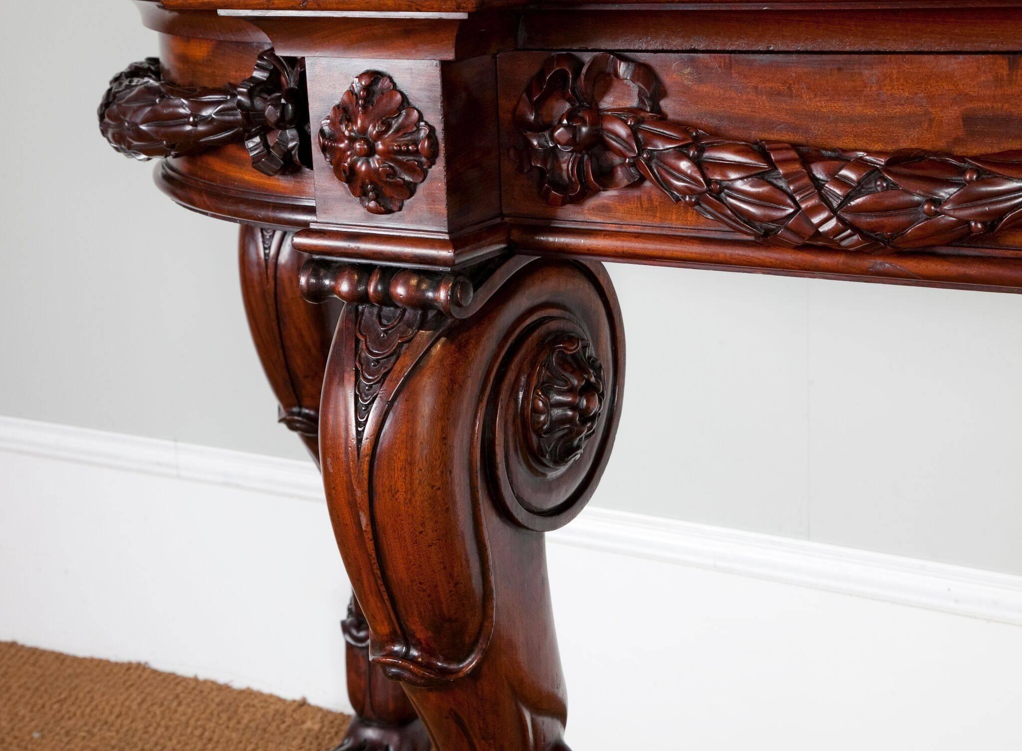 A robust George IV Irish console table. The rectangular mahogany top is enriched in color, with a moulded breakfront edge and bowed ends.
The top rests on a boldly carved frieze with laurel leaf swags, above four carved cabriole legs terminating in