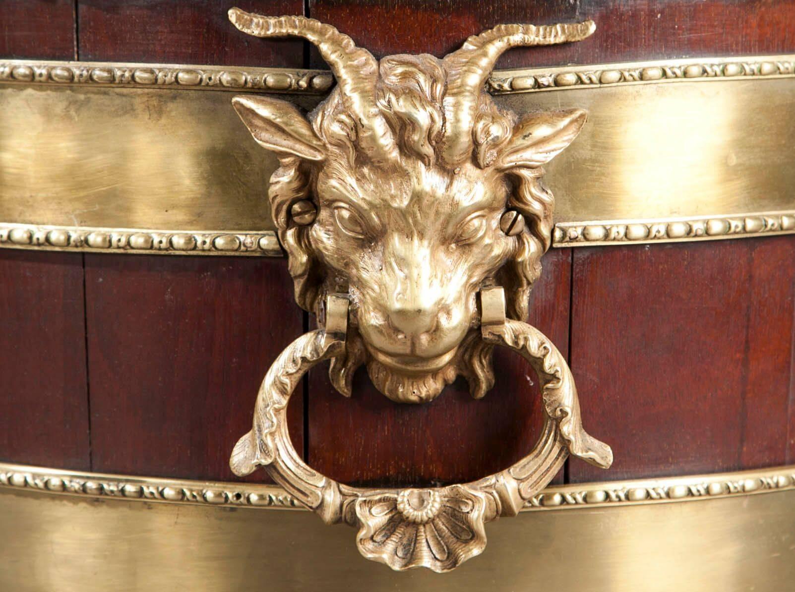 A George III style mahogany, ormolu-mounted oval wine cooler on stand. The oval cooler with a Gadrooned brass rim above two brass bands with ram's head carrying handles, resting on an ormolu-mounted base with Bacchus face flanked by vines on each