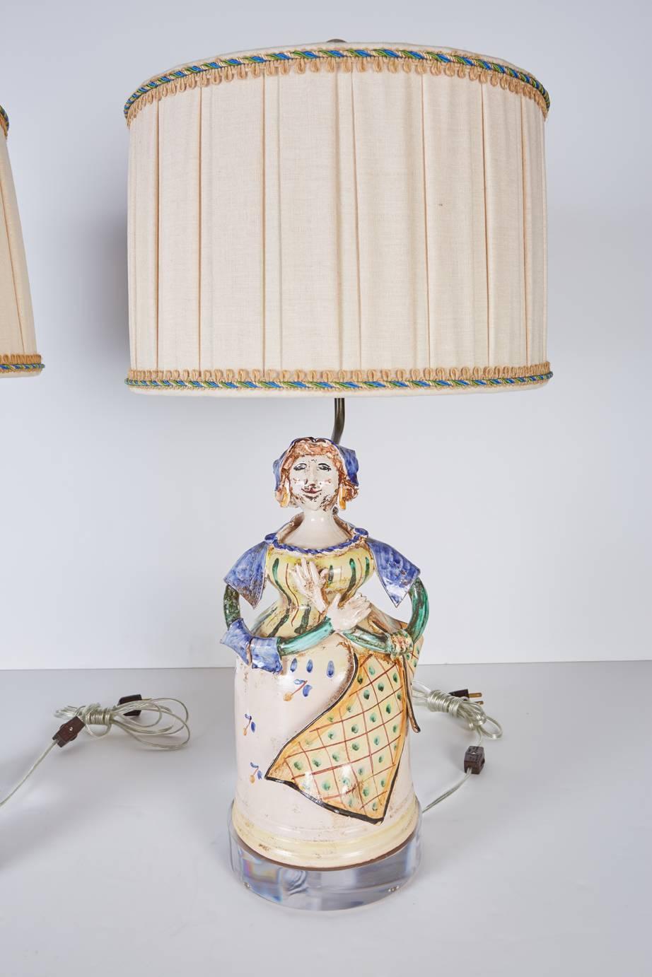 A pair of Italian midcentury figures mounted as lamps with custom Lucite bases and custom box pleated natural colored linen shades trimmed in twisted blue, green, and tan cording resting on a natural linen looped gimp.