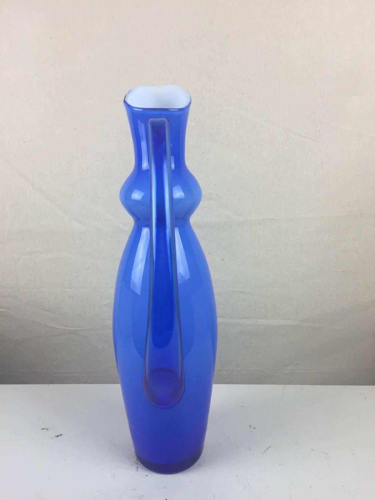 This graceful glass piece has a white glass interior and a brilliant periwinkle exterior and has stunning lines. It can serve as a pitcher or a vase. By Orrefors of Sweden. Signed on bottom. Width including handle is 6.5.