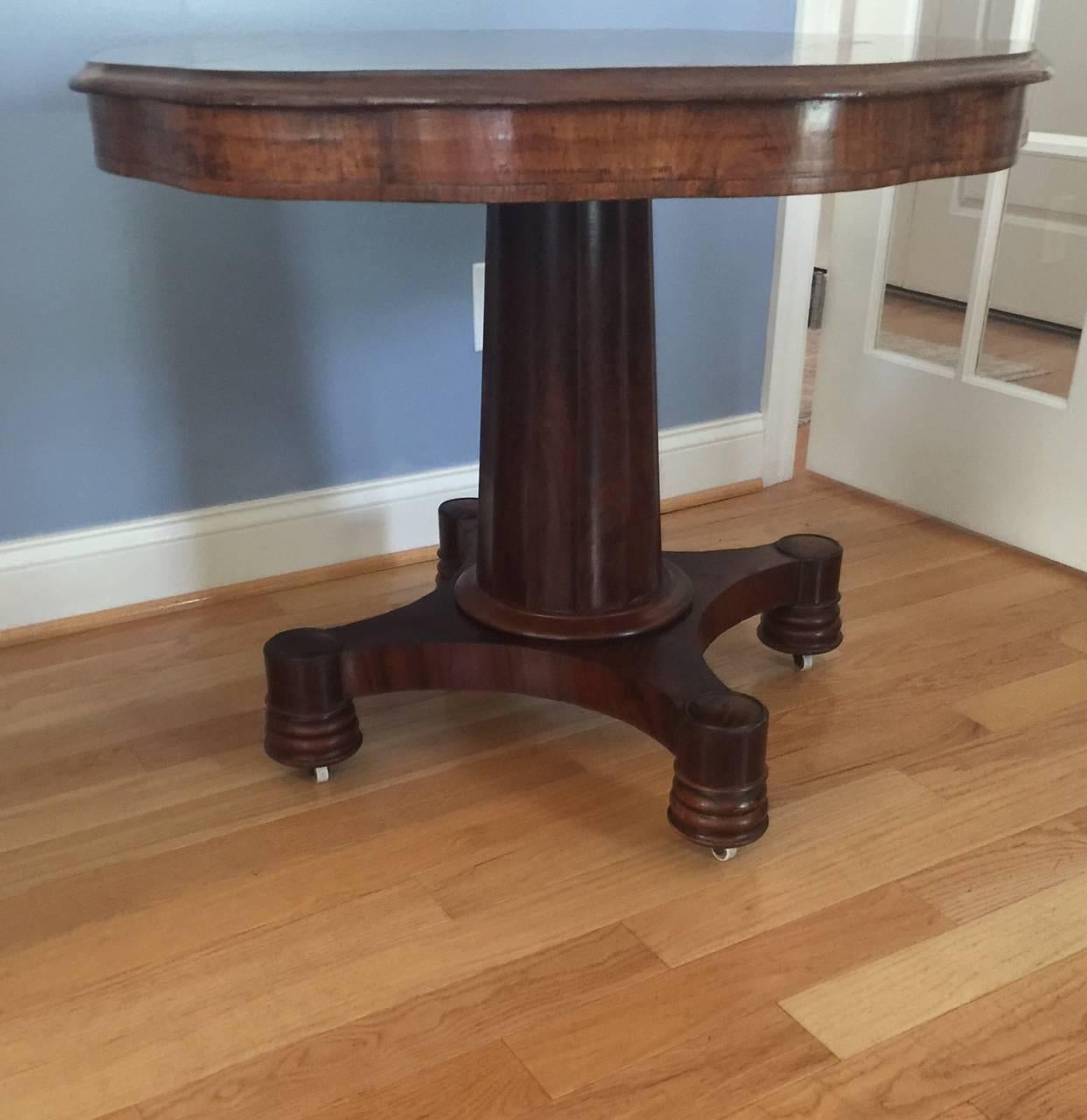 This handsome English piece has a flame mahogany top with beveled scalloped edge and apron. The pedestal is fluted. The base has a spool foot (and is set on wheels) with four legs. Although without markings, this piece is probably English Regency,
