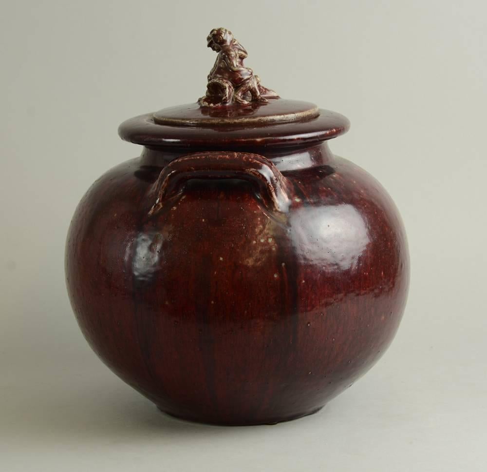 Stoneware jar with oxblood glaze and figure of a man and woman sitting on a log to lid, 1930s.