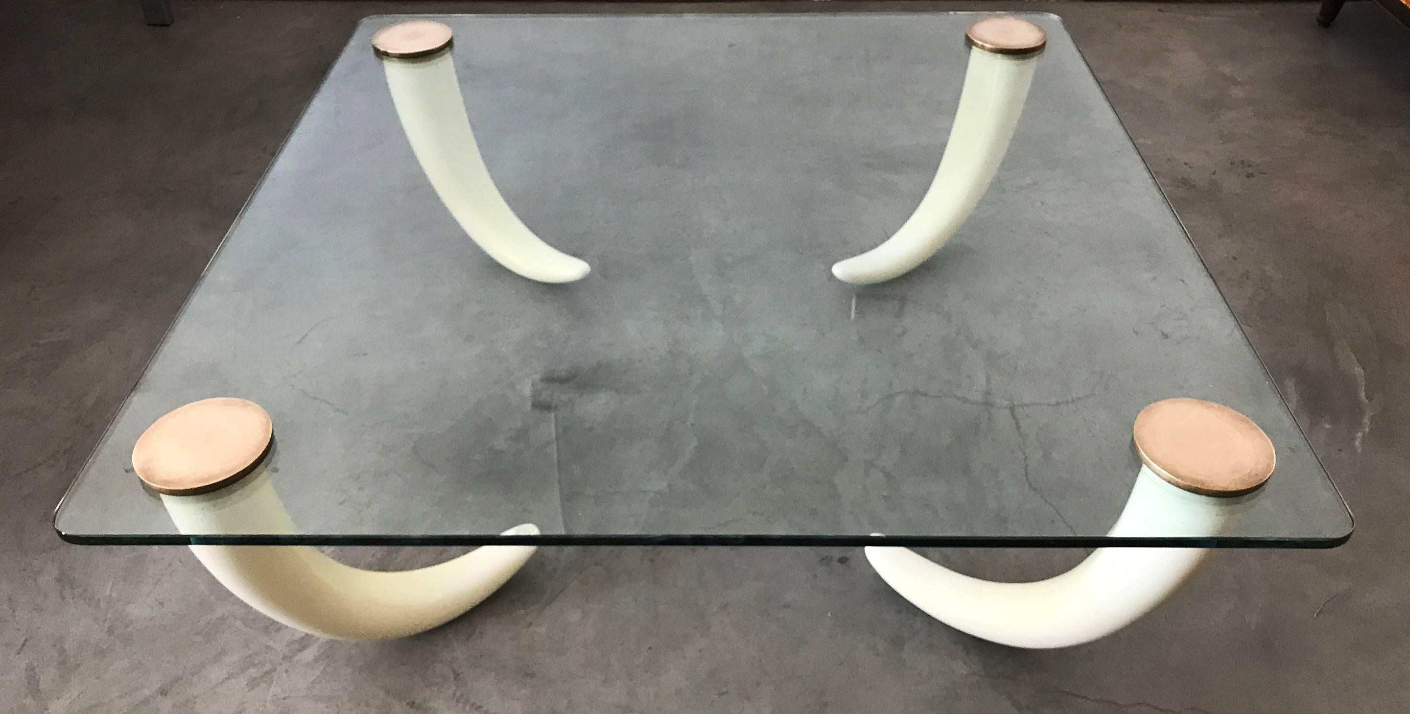 A stunning modern / Hollywood Regency style coffee table; this vintage, 1970s glass coffee table features four faux elephant tusk legs with solid brass caps which crew into the glass top.

 