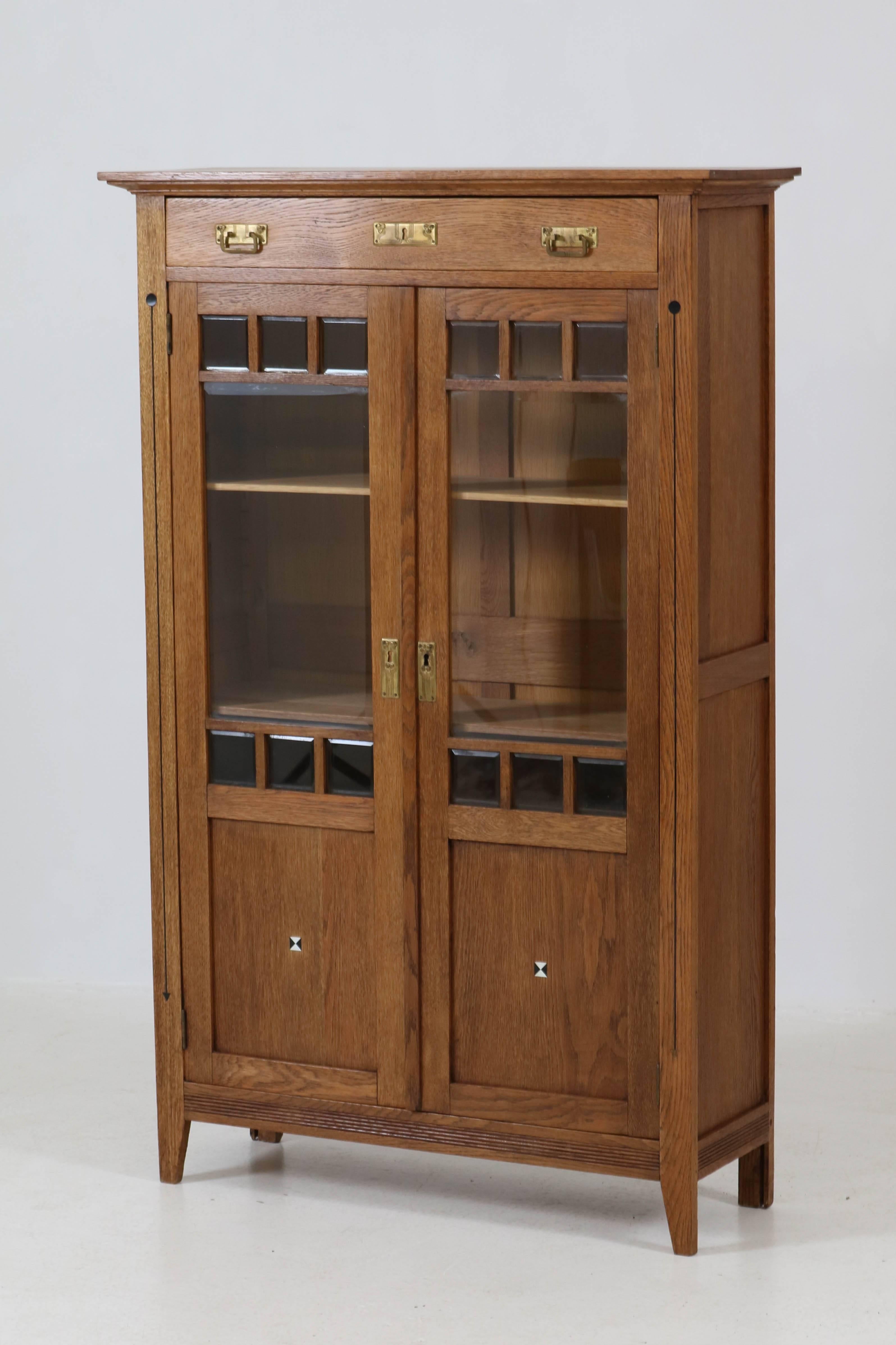 Stylish Art Nouveau Arts & Crafts bookcase with inlay, 1900s.
Solid oak with 14 original beveled pieces of glass.
Three original adjustable oak shelves.
Original brass hardware on doors and drawer.
In very good condition with minor wear