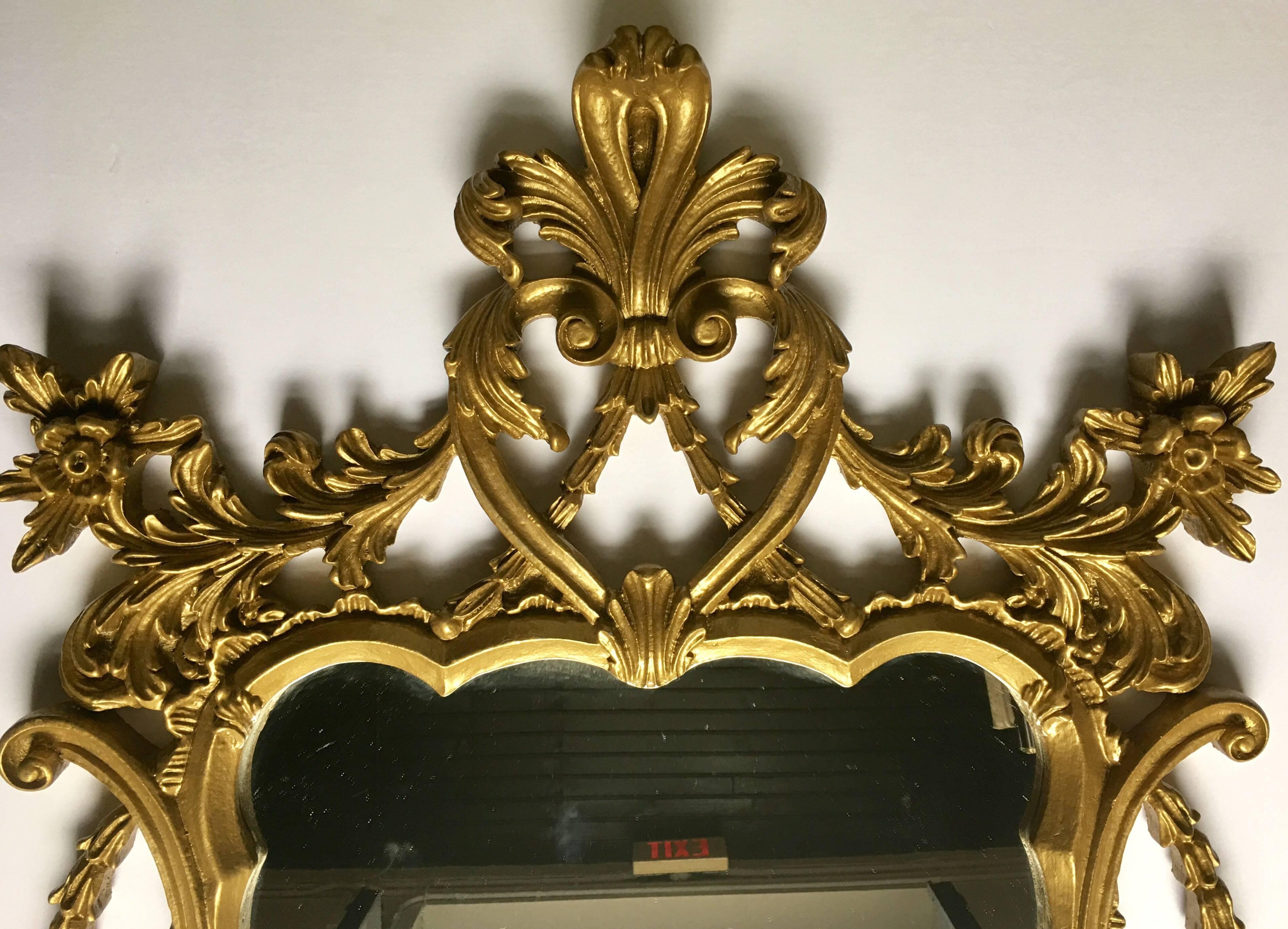 Midcentury molded resin/composition Rococo style wall mirror. This Hollywood Regency style framed mirror features a painted metallic gold finish with sculptural carved-like scrolls, acanthus draped garlands and leaves, and a fleur de lis cartouche.