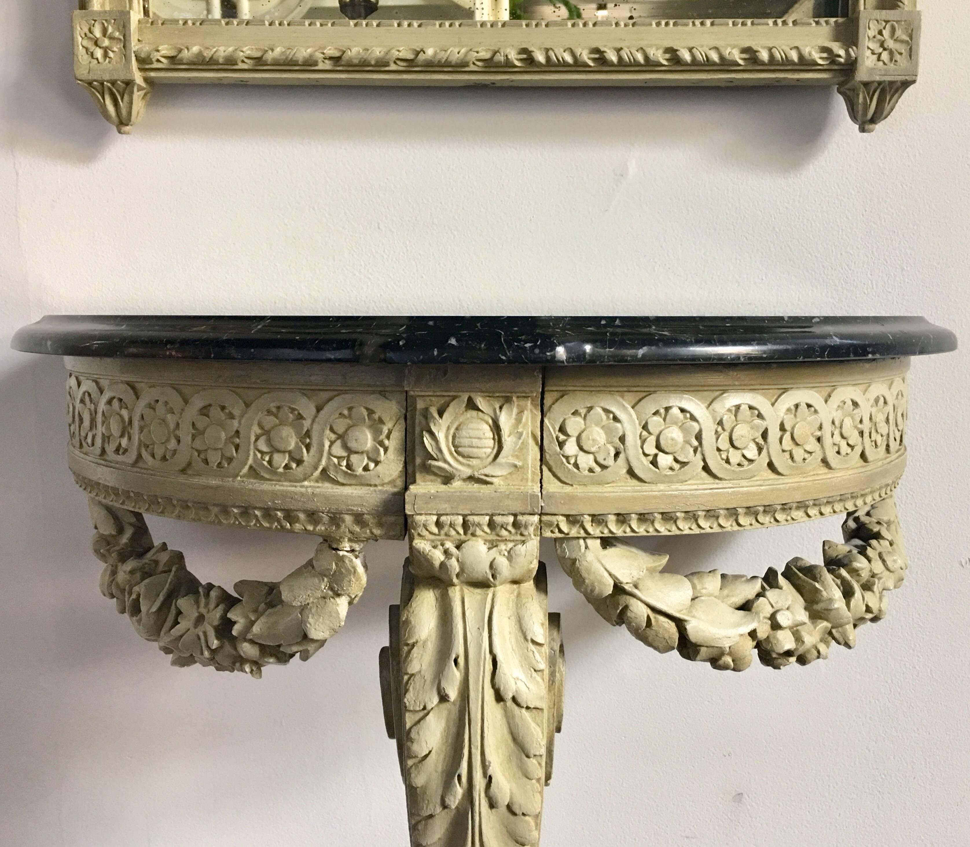 Louis XVI French style wall mount demilune console table and tall pier mirror. This small petite console table features single scrolling support and carved garland detailing with a new black stone top. Both pieces feature original cream tone painted