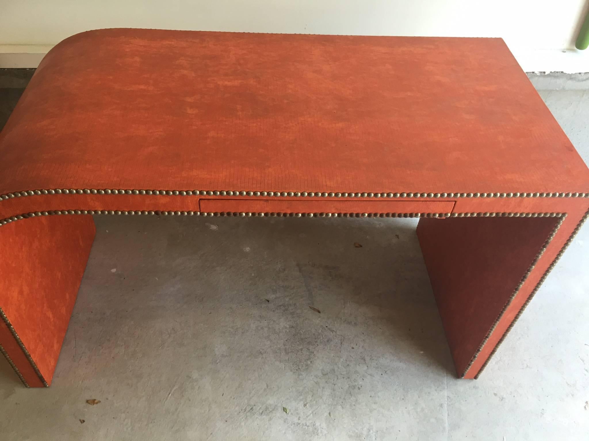 Rare Mid-Century Modern leather wrapped wood desk or console table. Sculptural design features original coral/orange embossed faux snake skin leather, aged nailhead trim detailing on front and back, and a single center drawer. Perfect size for your