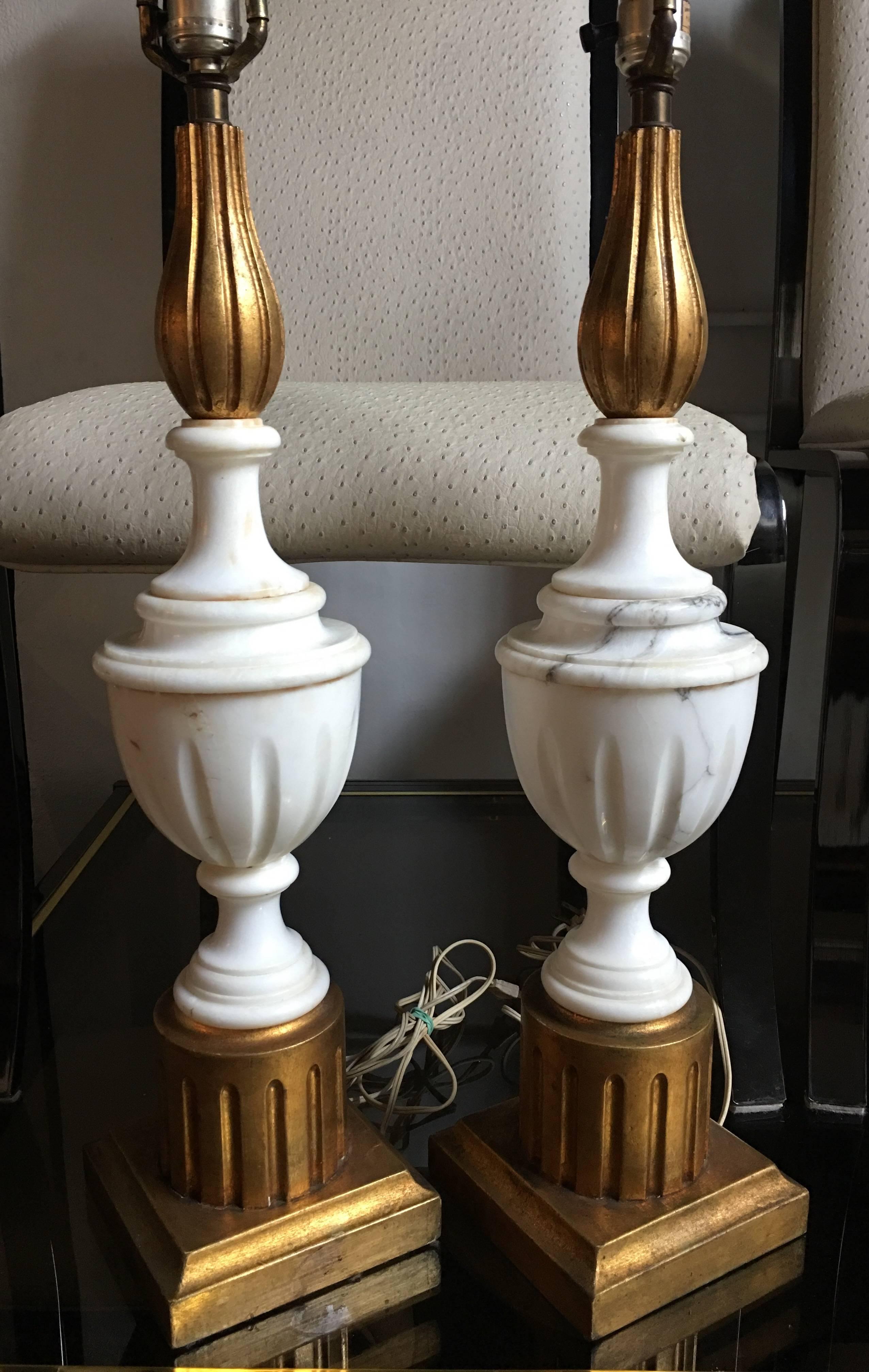 Pair of large Hollywood Regency style marble and gilt painted wood table lamps. Bases feature marble carved urns mounted on fluted giltwood column plinths.  One lamp has original 