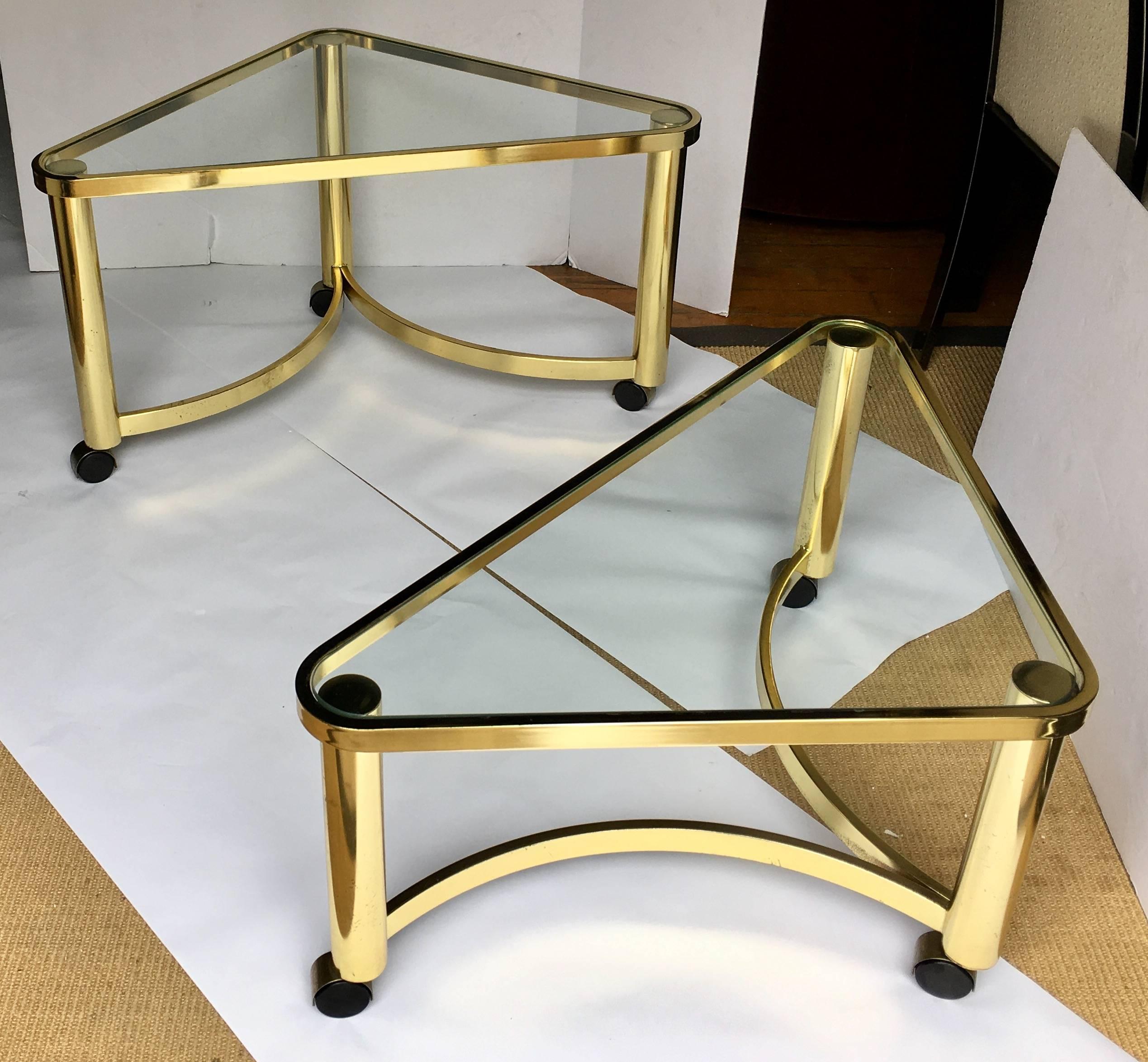 Set of two brass-plated sculptural nesting coffee tables by Design Institute America, DIA. These Mid-Century Modern tables feature clear glass tops with tubular triangular bases on rolling casters. These geometric tables can also be used as side or