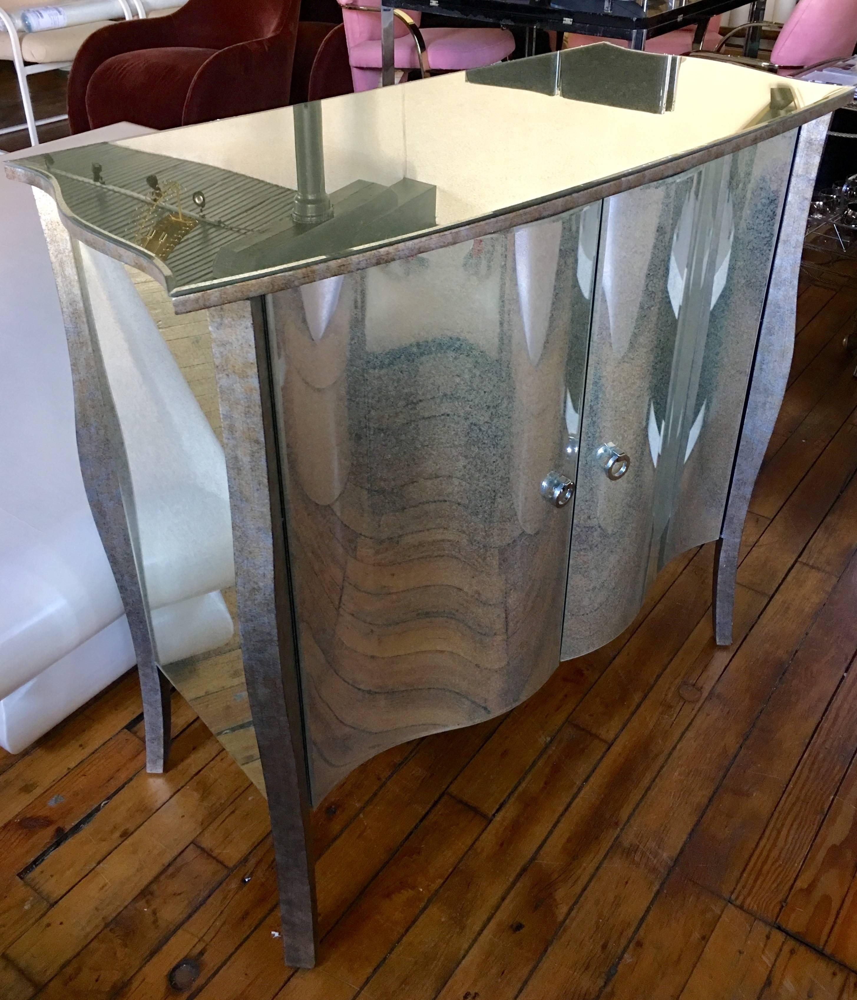 Glamorous Hollywood Regency style antiqued aged mirrored glass two-door chest featuring Polychromed wood, nickeled brass hardware and sculptural curved front doors. This versatile piece could be used as a bar cabinet, hall chest or dining sideboard.