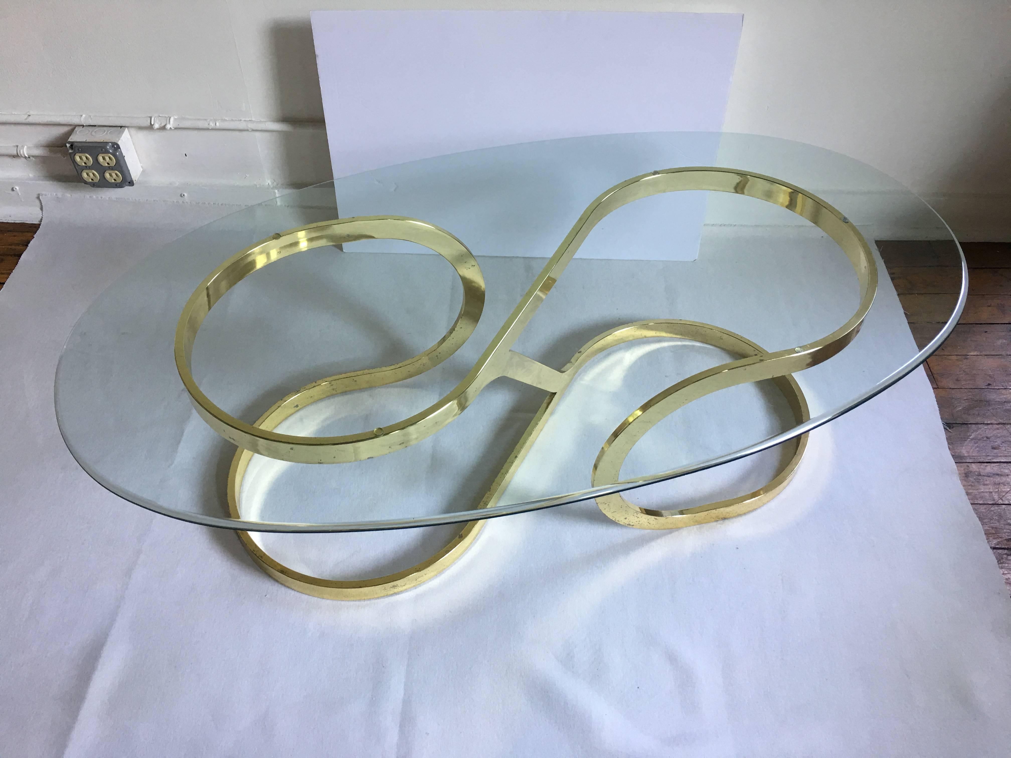 Large Mid-Century Modern glass and brass plated metal coffee table featuring an abstract curved twist design. Removable oval clear glass top. In the style of Milo Baughman. 
