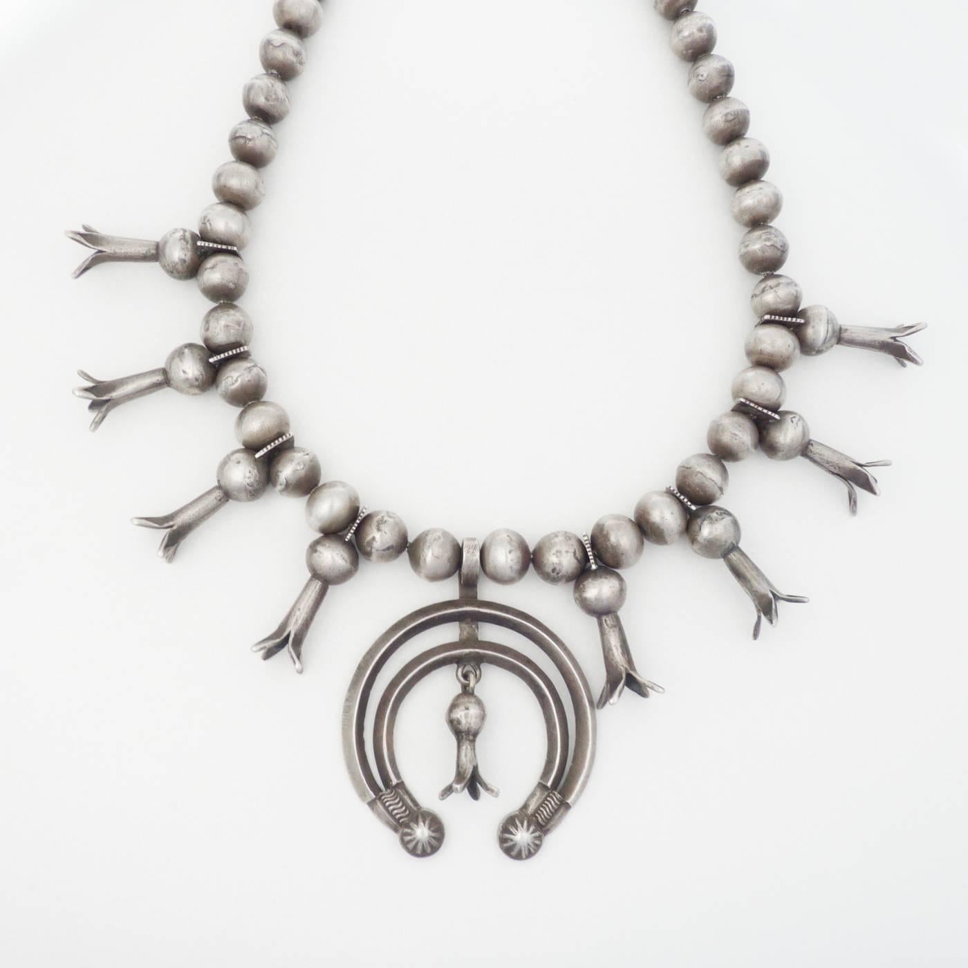 A Classic example of a silver Navajo squash blossom necklace, with an unusual naja that was cast and then had buttons applied to the terminals. Handmade round and squash blossom beads, and finished with a native wrap. Weighs 257 grams, is 27 inches