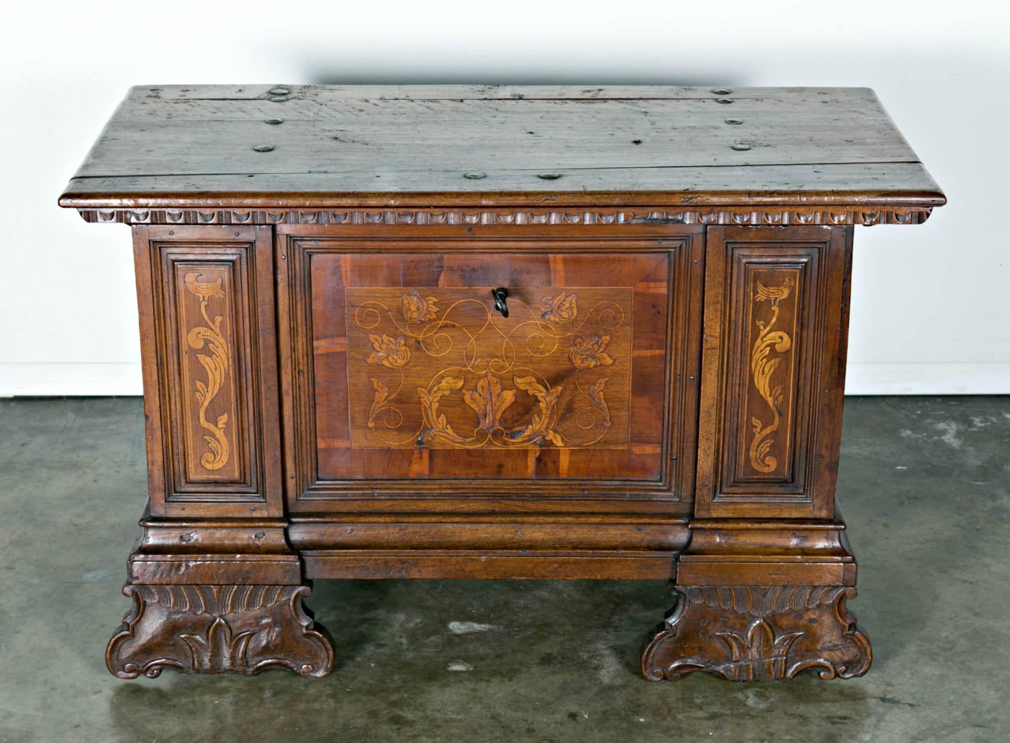This rare, rectangular Italian cassone or trunk from the late 1600s is of solid carved walnut with intarsia-inlaid, string banding, having hand-forged iron fittings. The trunk rests upon two carved s-scroll feet and two back block feet. Original