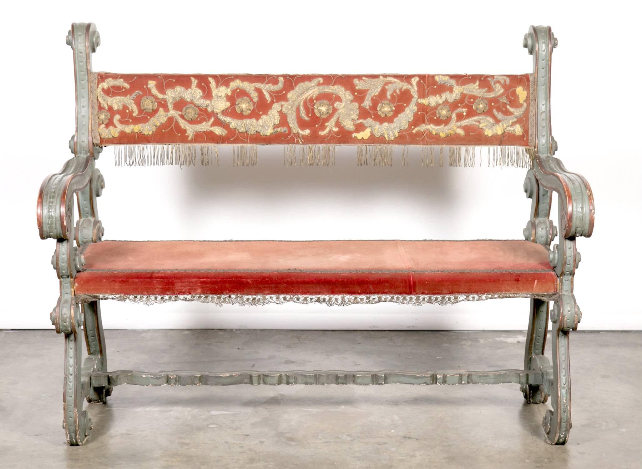 A fabulous pair of 18th century painted Baroque style Tuscan arm benches with carved lyre shaped legs joined by a carved stretcher and upholstered with antique ecclesiastical embroidered fabric and fringe. The well defined and raised metallic gold