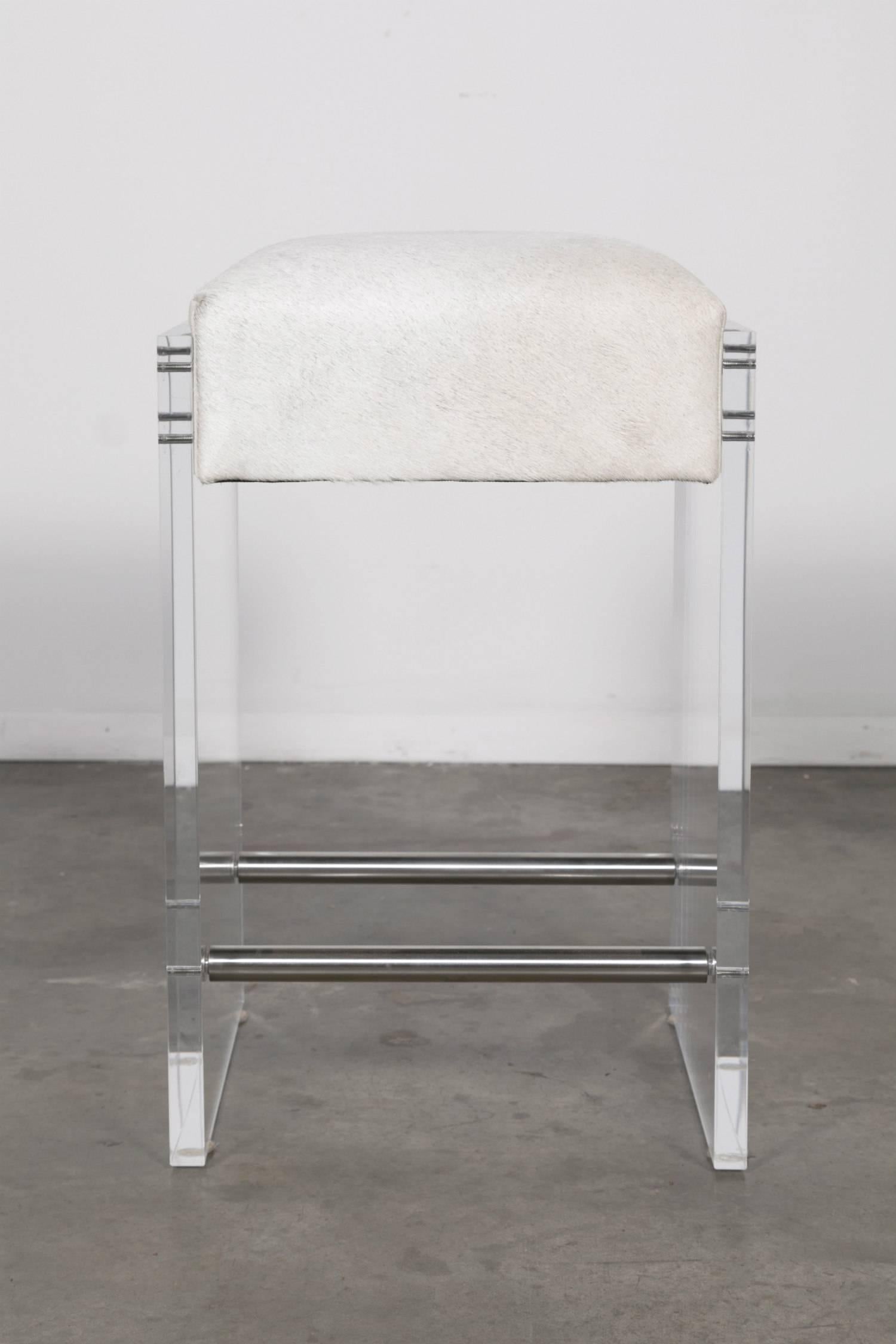 Gigi Lucite counter stool with two stainless foot rests. Add style and a touch of Hollywood Glam to your decor. Modern acrylic frame fastens to cowhide seat featuring cream faux leather sides with polished stainless steel fasteners. Counter stool
