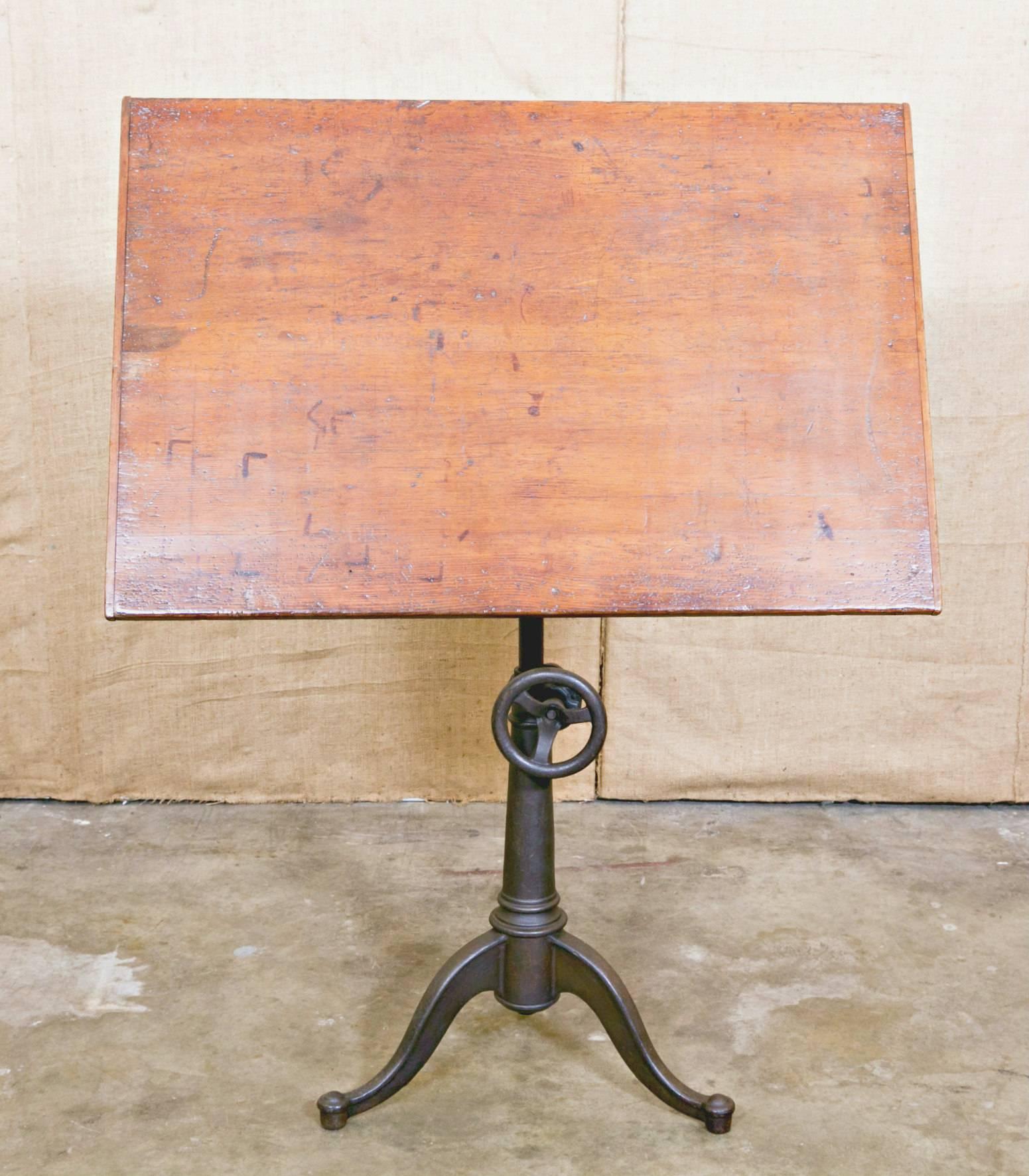 Vintage American drafting table with cast iron articulating tripod base, having Industrial knobs allowing for the adjustment of table top height and tilt. Original pine top measuring 25.5