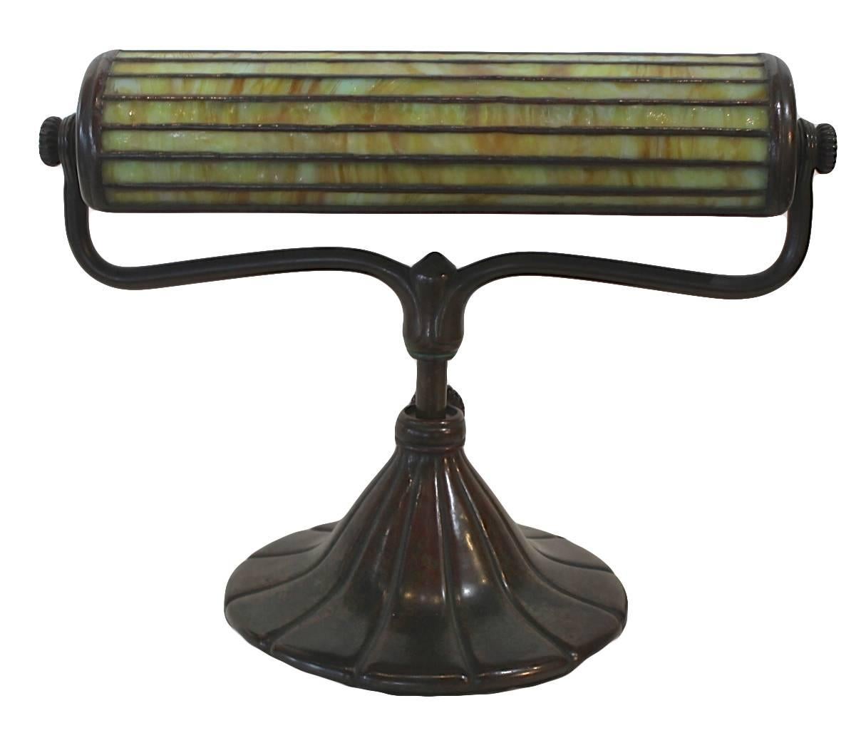 The cylindrical shade with horizontal panels of mottled pale green, white and gold glass symmetrically leaded, pivoting on a harp form bronze support and gadrooned circular base. Impressed Tiffany Studios New York
Provenance: The John L.Severance
