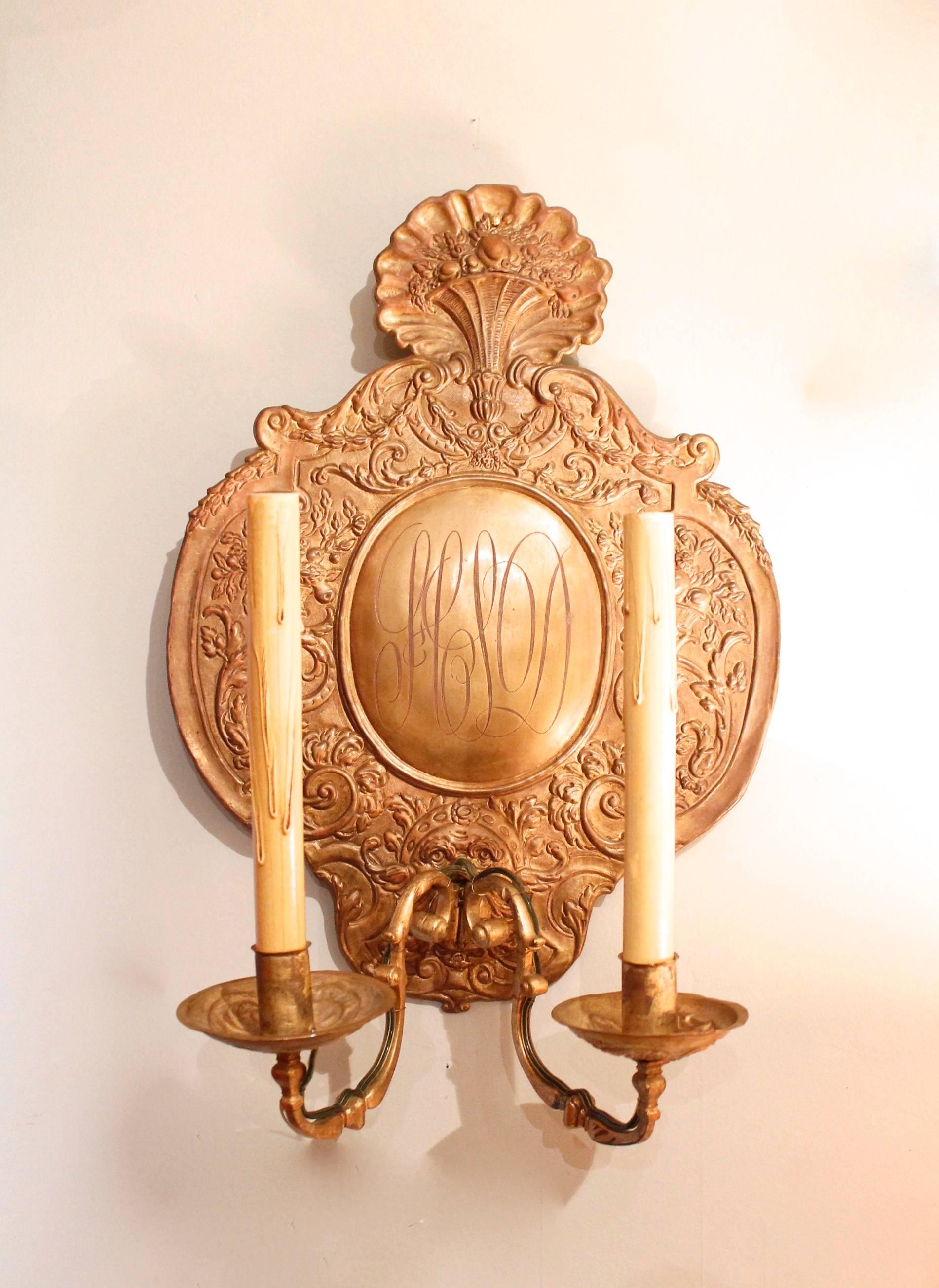 A pair of electrified repoussé brass sconces, embellished with foliate designs around oval center bosses which are engraved with the initials HSD.