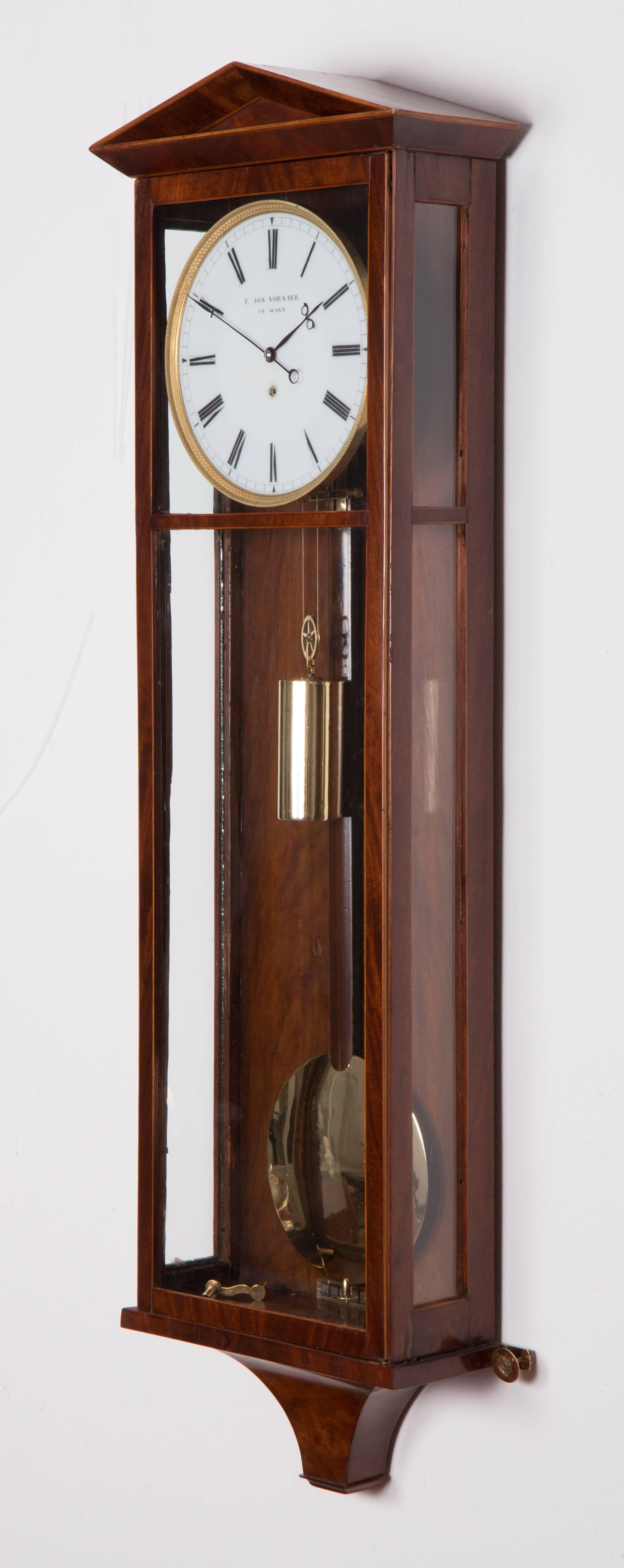 Measures: Length: 89 cm, duration: eight days
Mahogany veneered case with maple stringing, milk glass dial, blued steel hands, gilt and engine turned bezel, deadbeat escapement, spring suspension, wood rod pendulum. Stamp at the back plate: “F.