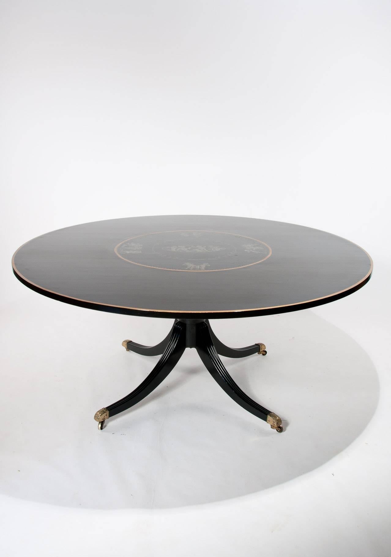 A large 5 1/2 feet round ebonised Hollywood Regency style dining table on central splay base circa 1970-1980s. 
The large 66-inch diameter top having a hand-painted central round of a woman surrounded by cupids enclosed by a gilt round edge