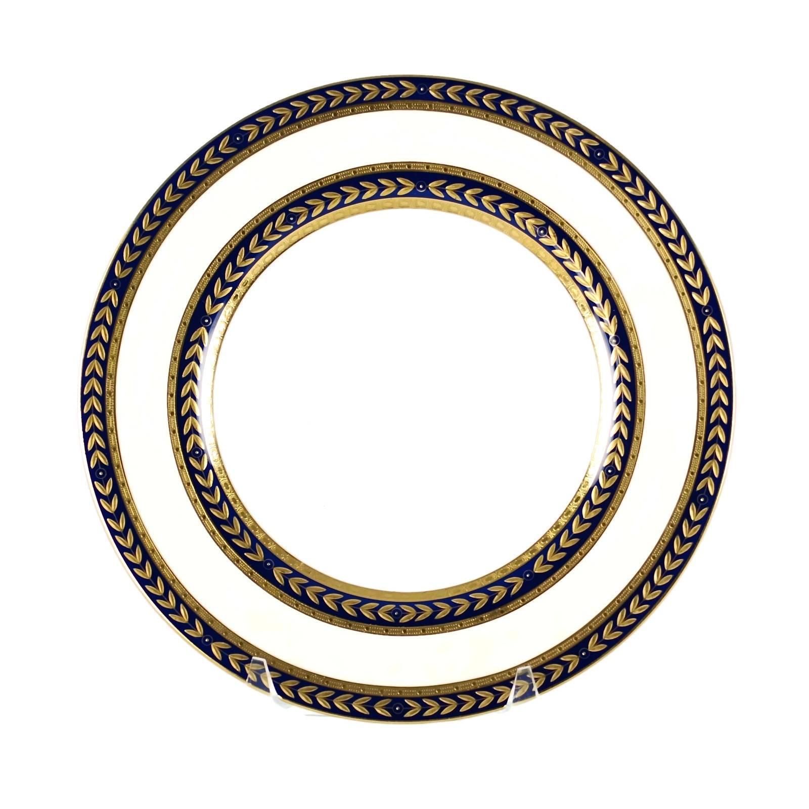 This elegant set of 12 porcelain service dinner plates was made in England by Mintons for retailer Tiffany & Co. The pieces have a white ground and feature cobalt blue bands bordered in gilt and accented with raised gilt laurel leaf decoration. The