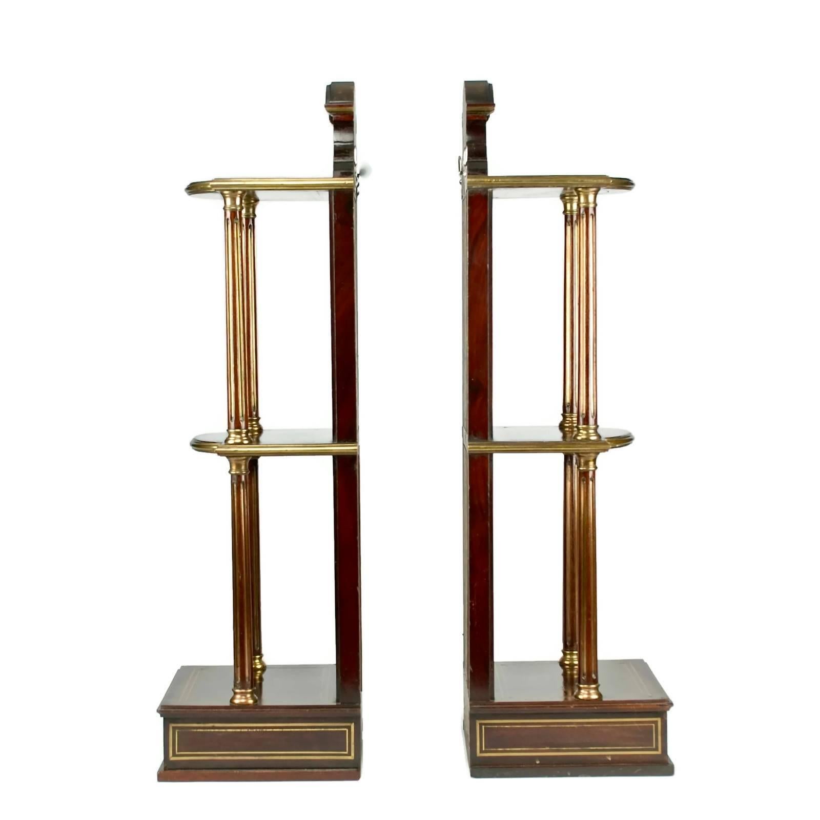 This handsome pair of French Empire mahogany wall shelves feature beveled mirrored backs and fluted columns. The shelves are trimmed with inlaid brass accents and have a single drawer at the bottom. The pair are fitted with hooks at the back for