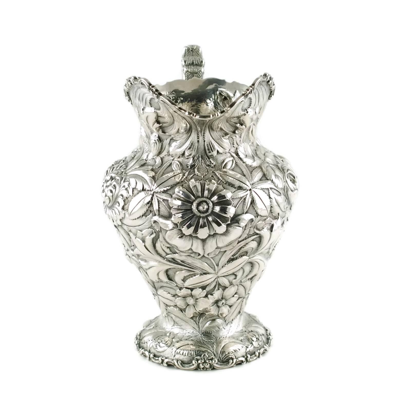 This large sterling silver water pitcher was made by the Stieff Company of Baltimore, Maryland. The hand chased piece has been finished in Stieff's ornate Repousse pattern and features lavish floral and foliate decoration on a stippled ground. As