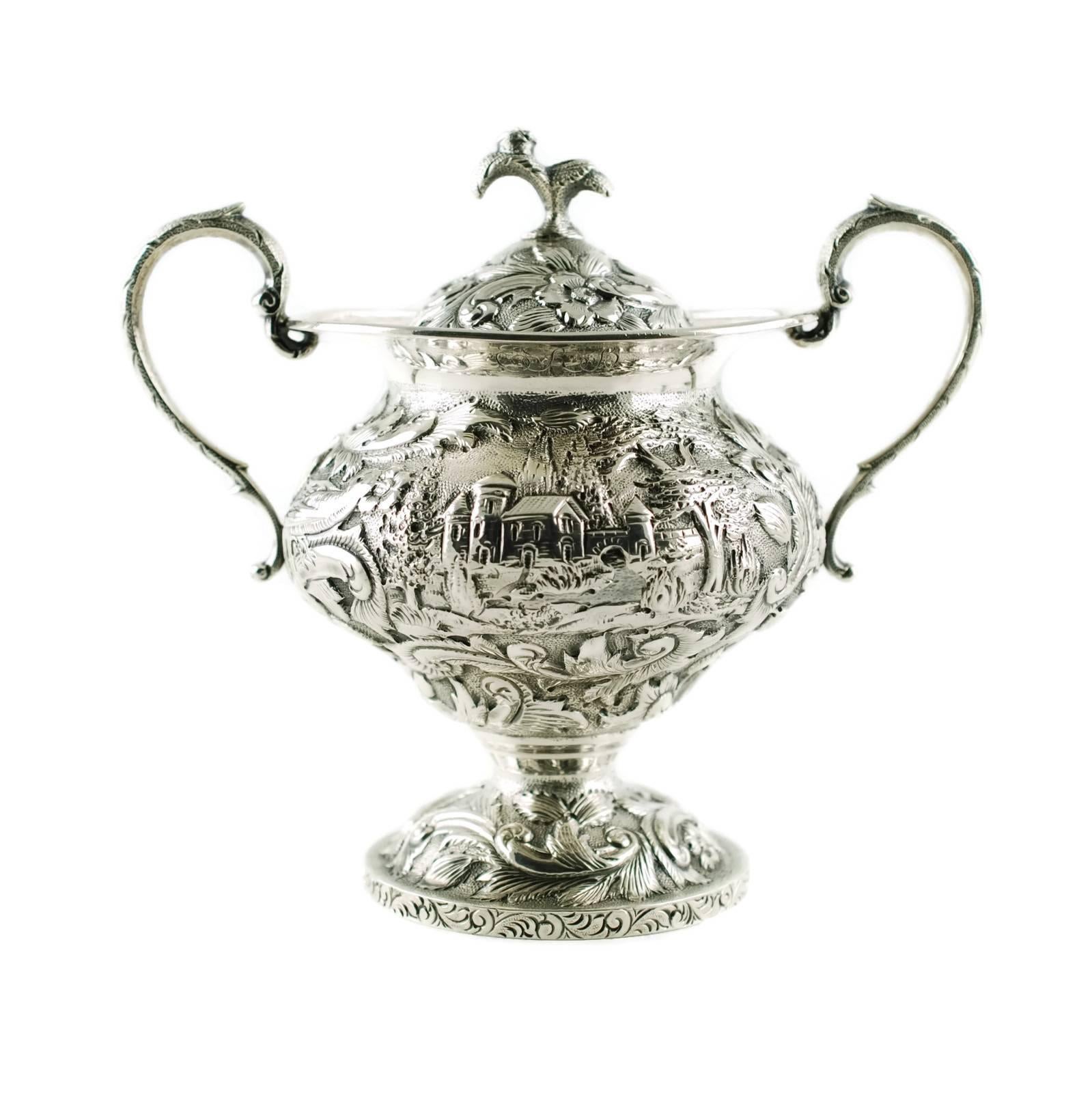 This early 20th century sterling silver three-piece tea set has been made in the ornate Castle pattern and was retailed by The Loring Andrews Company of Cincinnati. The set consists of a pedestal form teapot and matched covered sugar bowl and cream