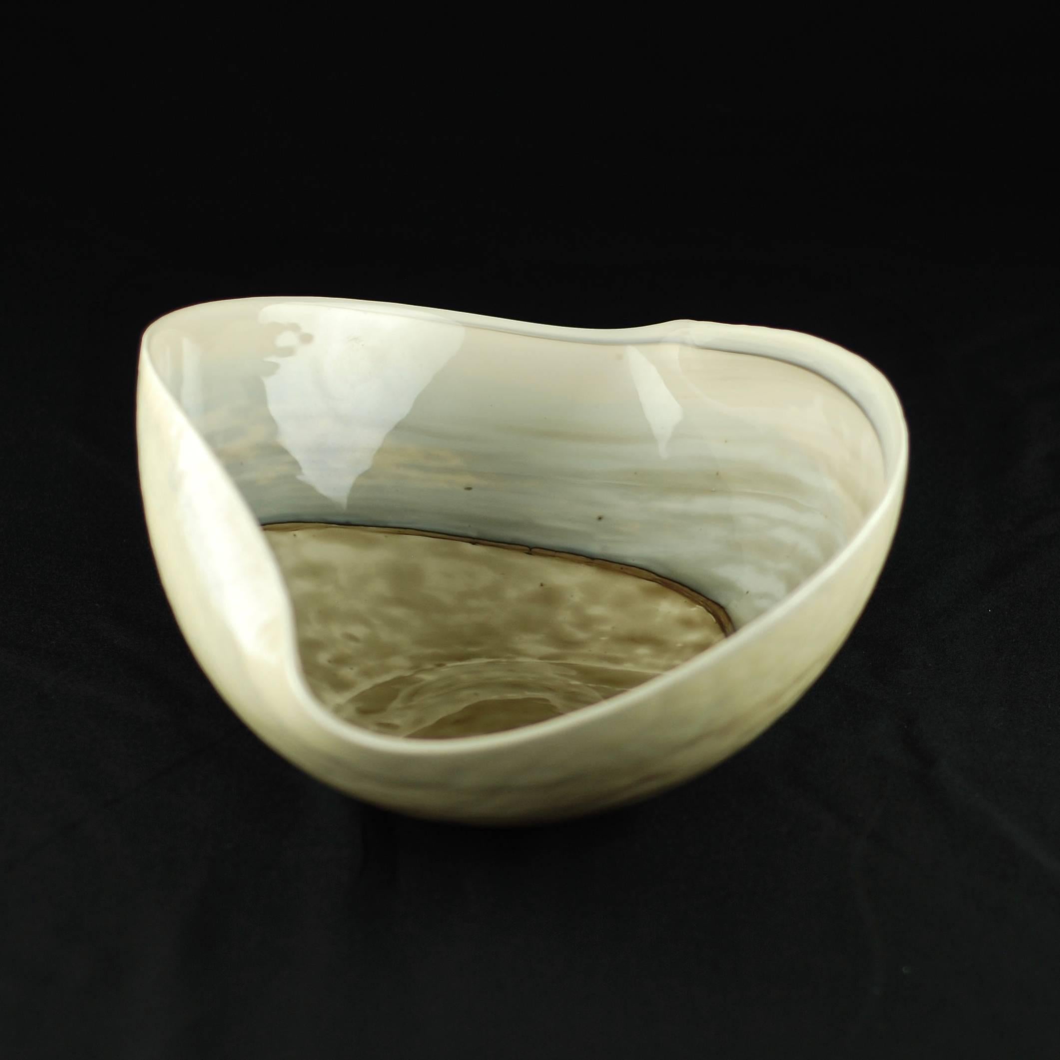 This large art glass centerpiece bowl has a freeform ovoid shape with folded sides and features an abstract swirling pattern. The piece has been executed in a neutral palette which includes shades of cream, tan, brown and blue/gray. The interior of