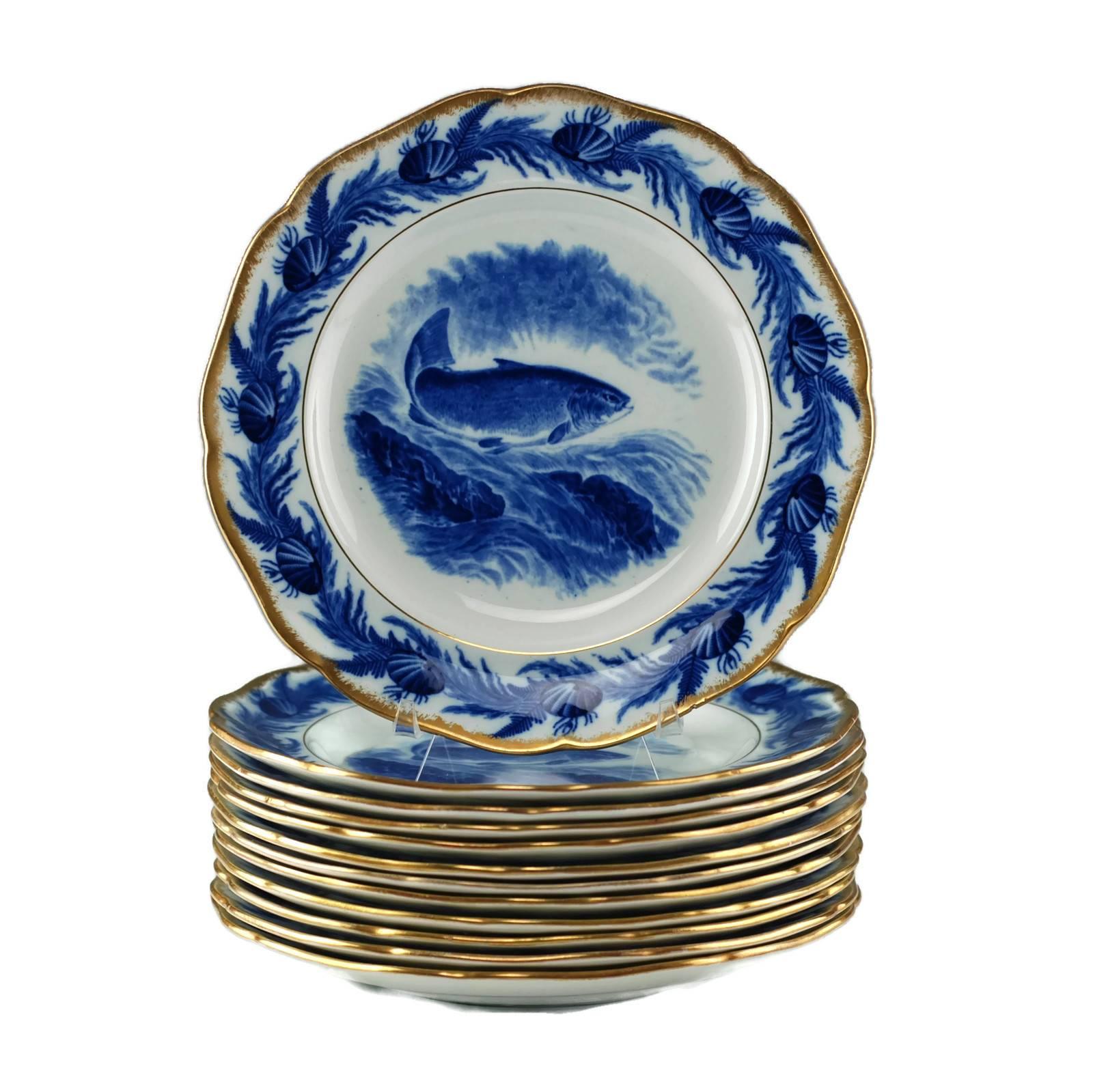 This set of 12 antique Staffordshire flow blue plates was made by Cauldon England and feature the image of a large salmon leaping above the surface of a stream which is cascading over rocks. The central image is encircled within a band of aquatic