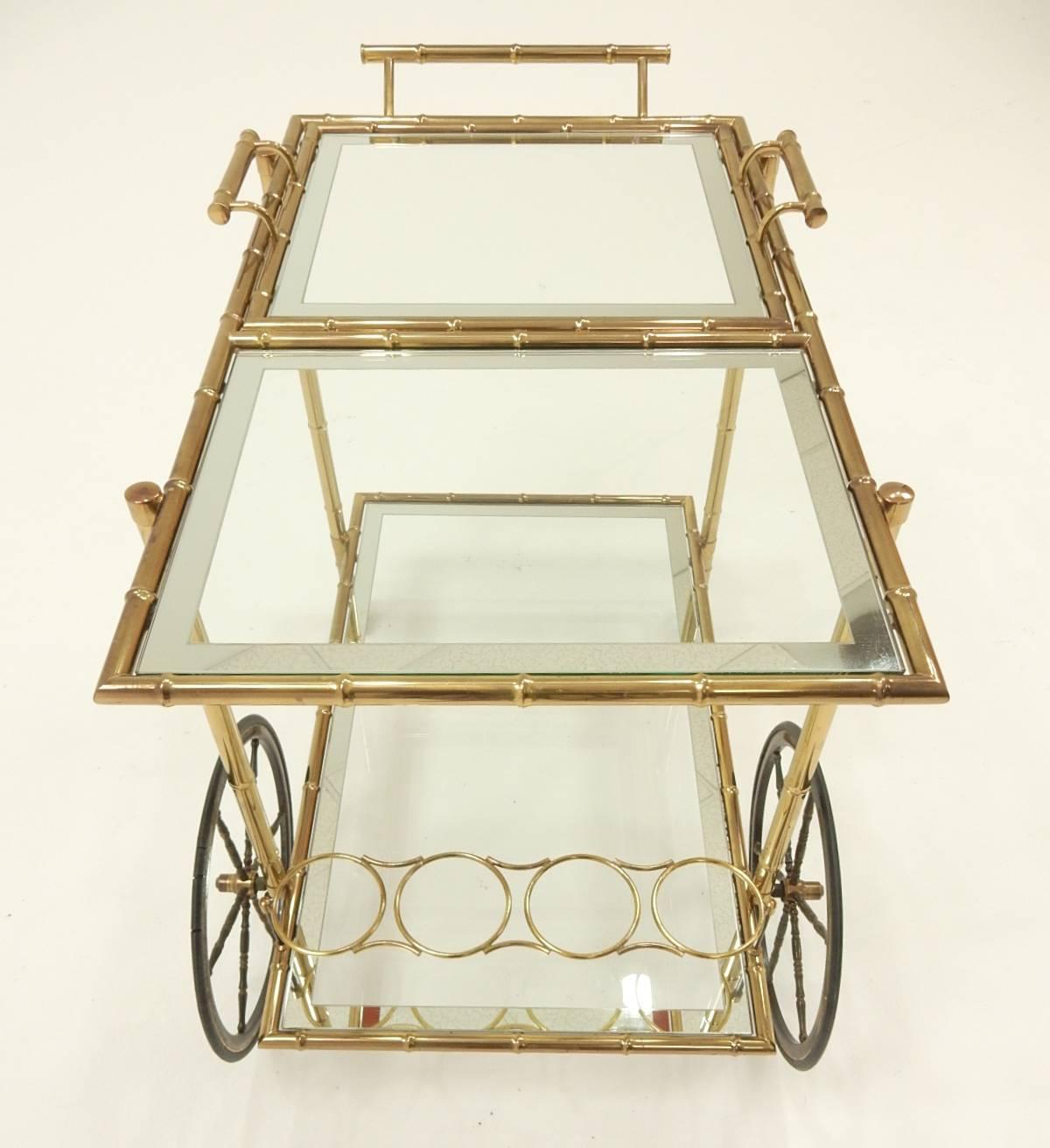 Brass faux bamboo bar cart with mirror edge glass, circa 1960s.
Three ring bottle/decanter holder in lower section.
Top half is removable tray with handles for serving prepared drinks.
Large spoke front wheel with smaller spoke rear