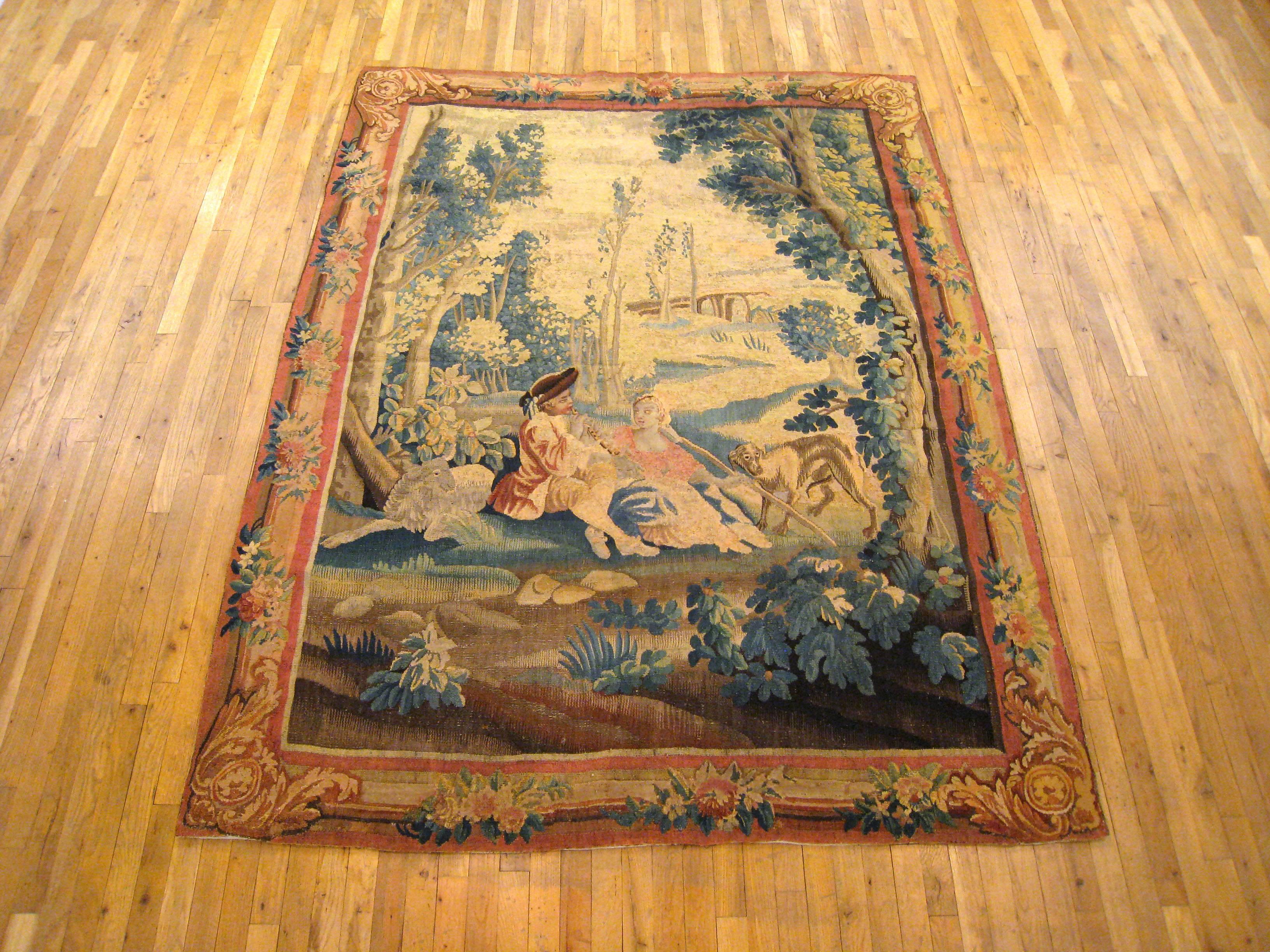 An antique 18th century French pastoral tapestry, size 8' H x 6' W. This lovely tapestry portrays a tranquil scene, in which a gentleman plays flute for his dame while they repose in an idyllic woodland setting, with their dog and lamb nearby.