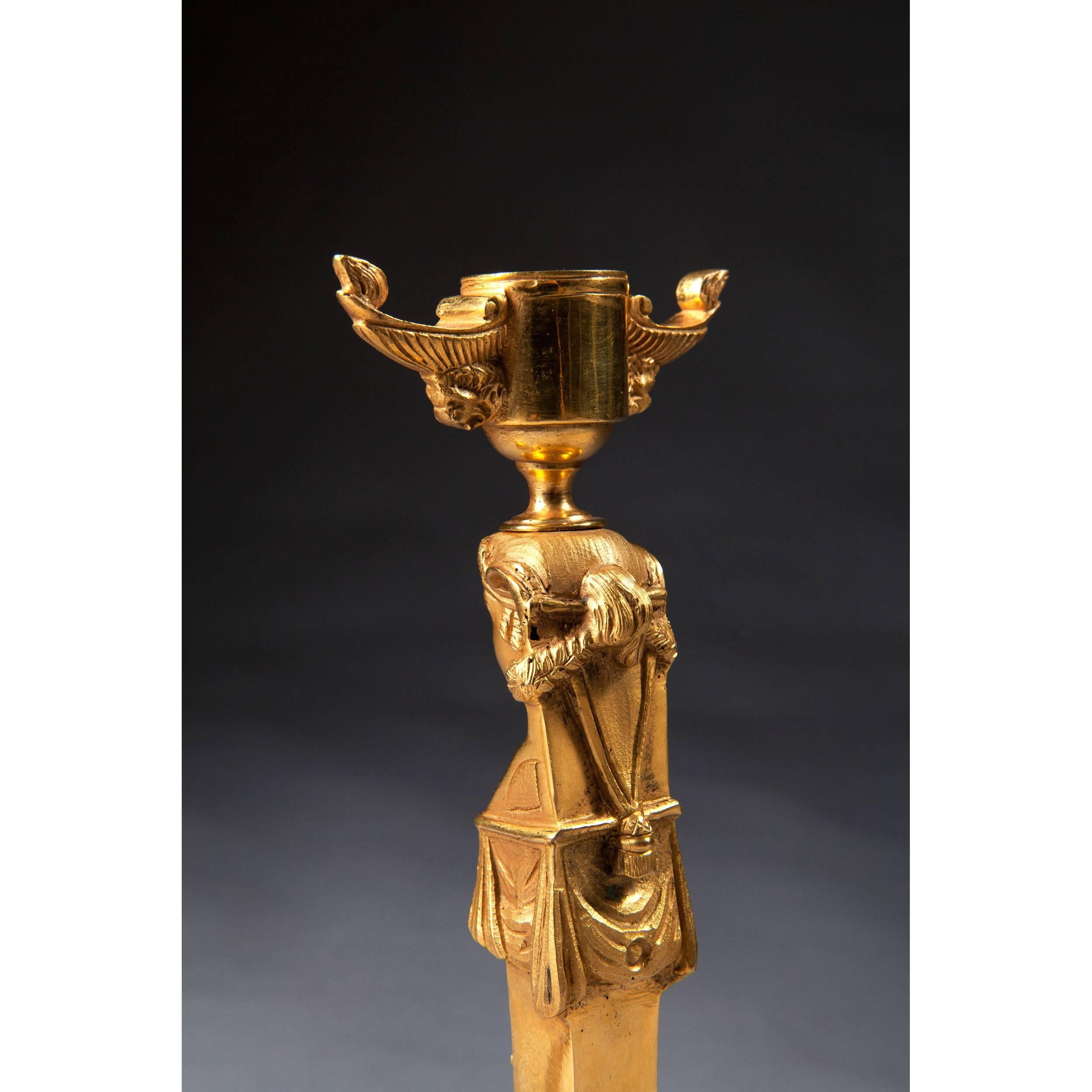 A fine pair of gilt bronze candlesticks as standing female Egyptian caryatid figures surmounted with gilt bronze handled nozzles with lion masks, the candlesticks of tapering form, the front mounted with vase candelabra detailing, each on circular