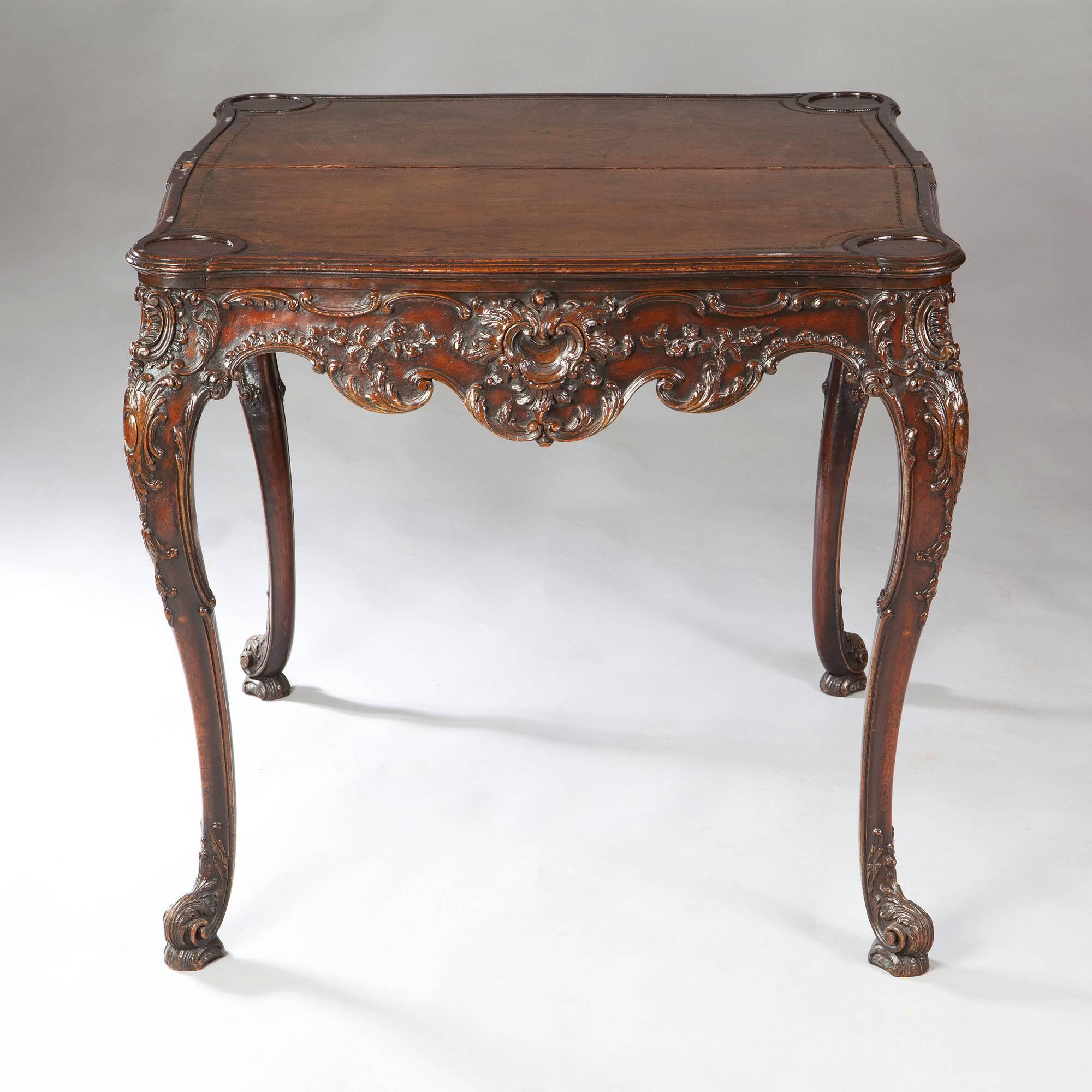 England, 19th century.

A fine rococo card table, the scalloped shaped front richly carved with rococo ornament, the flip top supported on extending rear legs and opening to a leather playing surface with four counter wells. The whole raised on