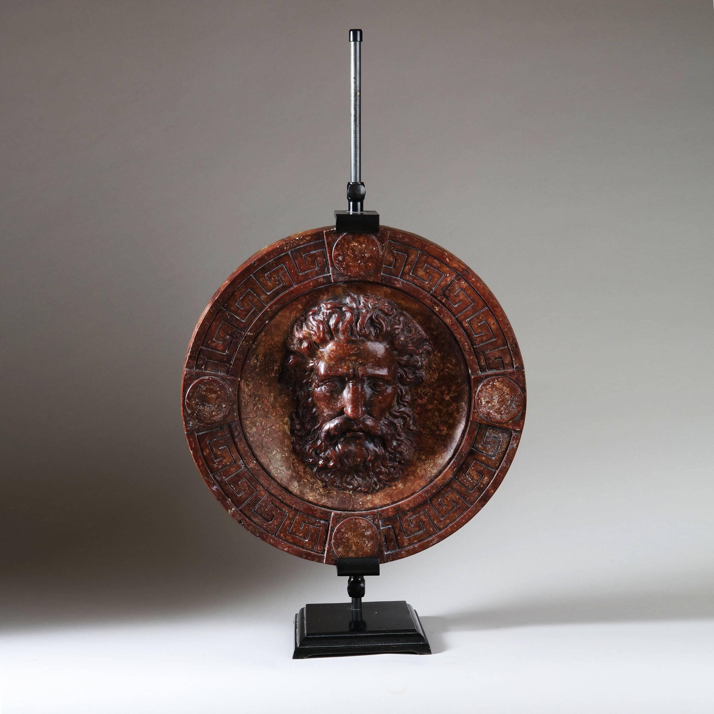 An extremely well-made copy of a classical mask after the antique, the face shown in high relief within a dished background and a border of Greek Key with discs to the four sides. Now mounted on an adjustable metal stand.