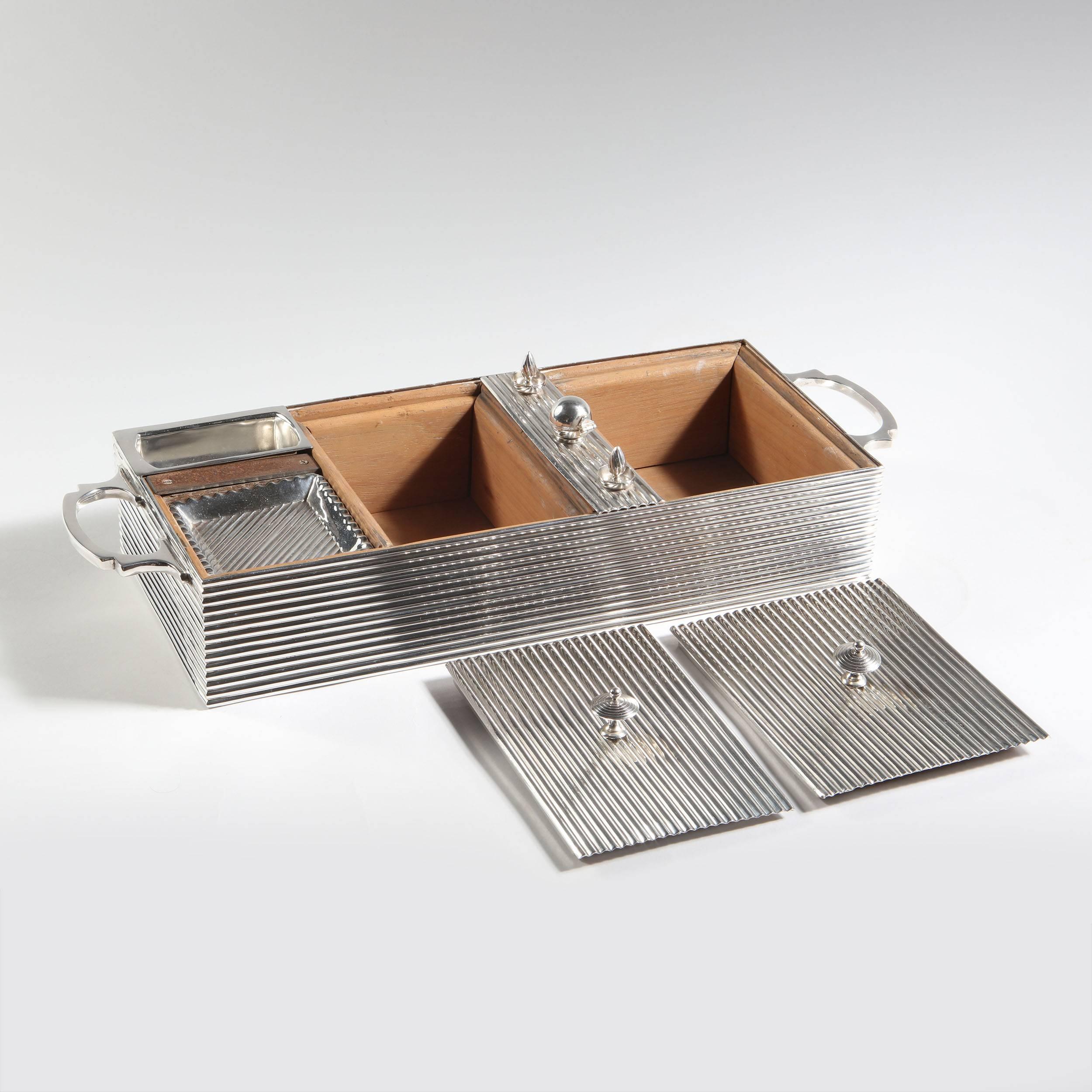 An unusual and inventive vintage silver box made to house cigars, ashtrays, a petrol lighter, two silver wicks and matches, each in their own separate compartment. The exterior in reeded silver plate with two handles, the two lidded cigar