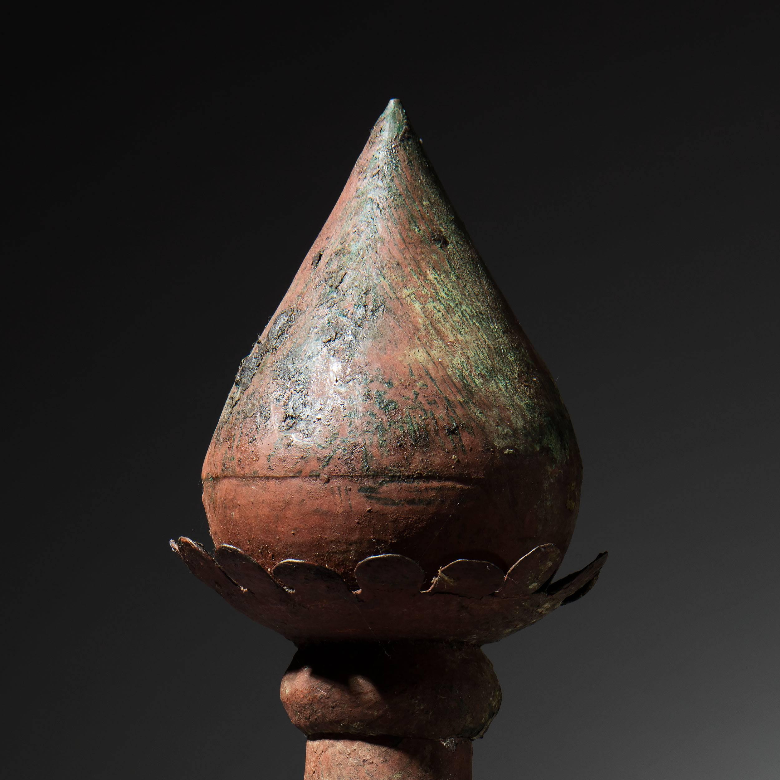 An unusual Indian tole finial painted a rust hue with touches of green over painting, inspired by a flower bud, a spike rises high above the base with stylised leaves and petals around two graduated spheroids, the whole on a flat oval base.