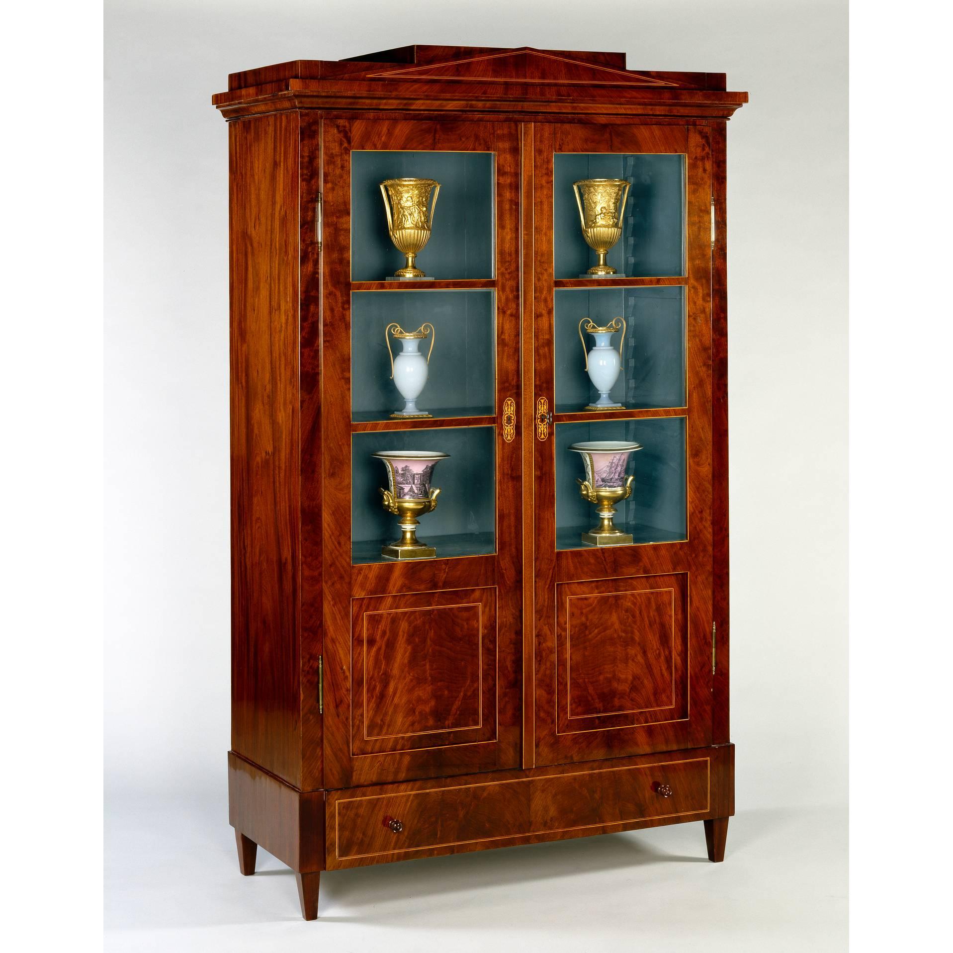 Germany, circa 1825.

 A Berlin mahogany display cabinet having a stepped pediment inlaid with boxwood stringing above partially glazed doors divided into four sections with scroll marquetry escutcheons.

Measures: Width 46in (117 cm),
height