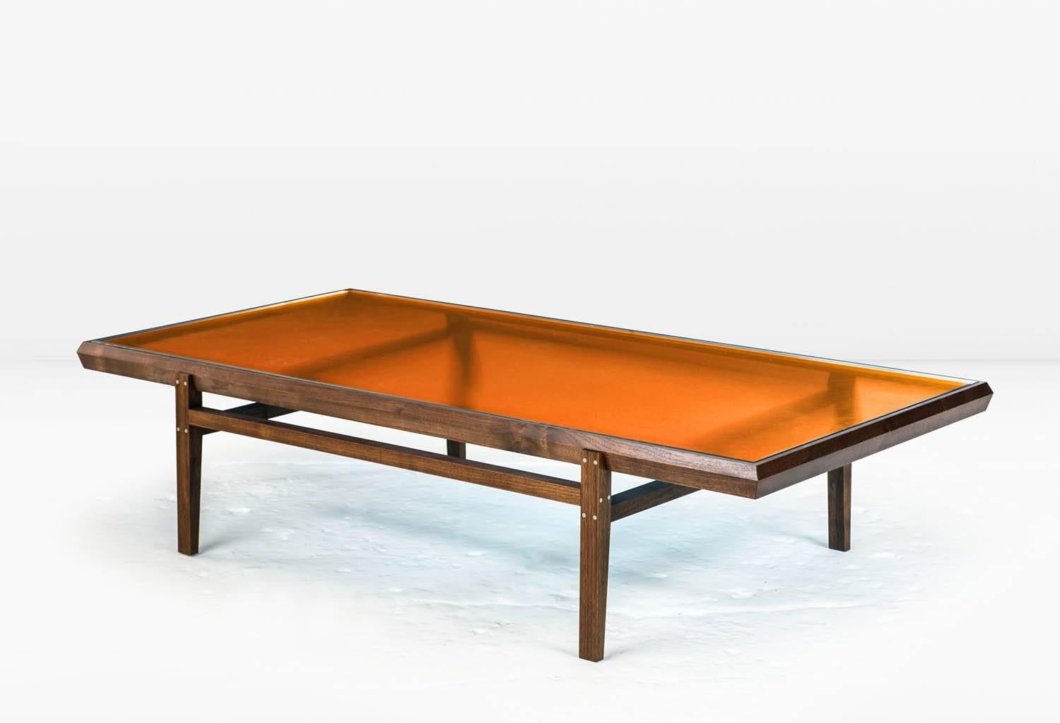 The Pintor coffee table’s solid wood frame with stainless steel inlay is engineered with chamfered edges to provide a simple but worthy setting for the jewel toned, colored glass which forms the tabletop. Shown with solid American black walnut frame