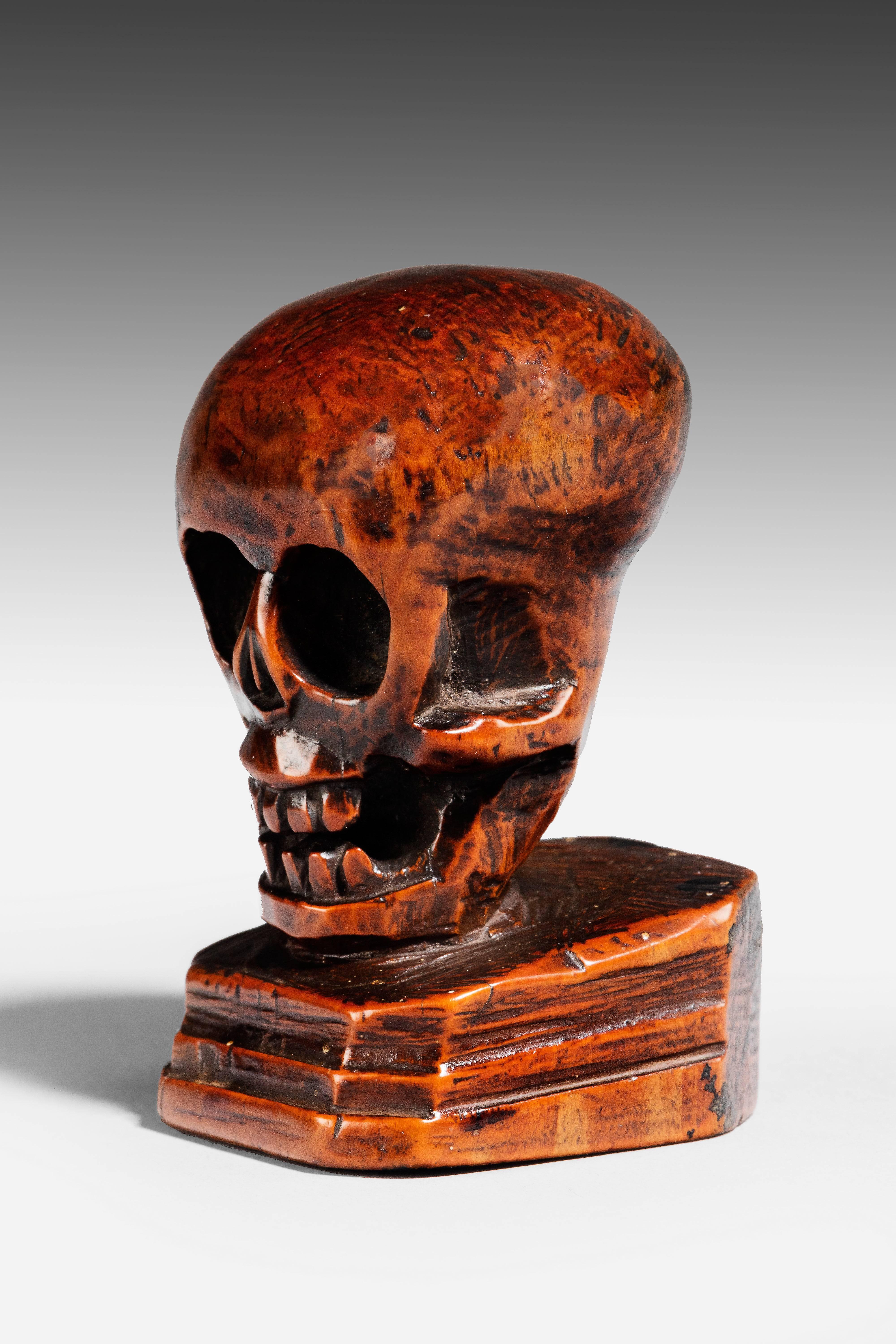 A well-patinated carving of a skull in boxwood. Modelled with gaping eye sockets and gaps between the teeth. Set on a sloping, stepped octagonal base. A ‘memento mori’ – an object designed to remind the viewer of their mortality and of the shortness