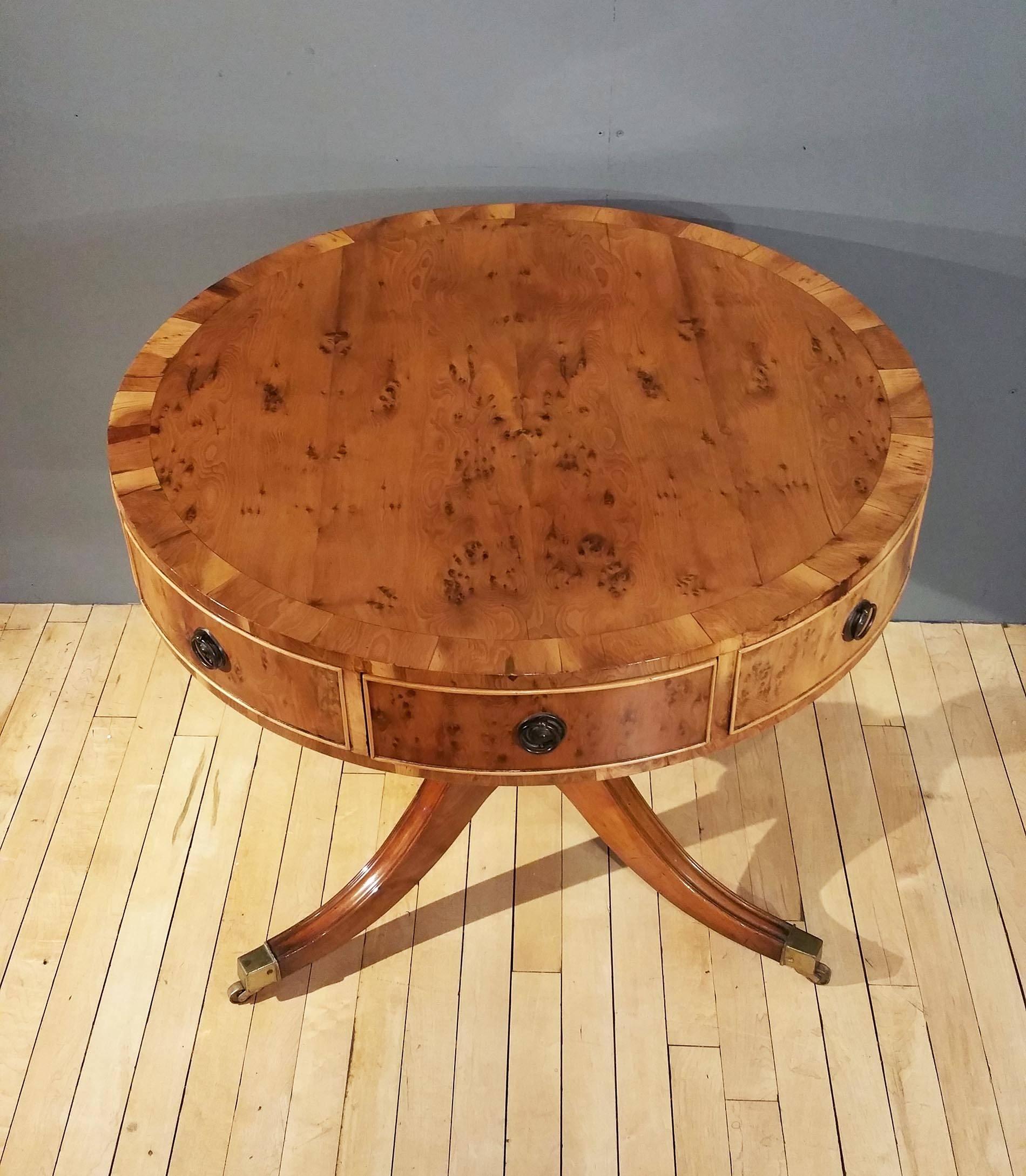 This lovely and fine quality yew wood mid-20th century drum table features alternating four display and four opening drawers on a central tripod base with sweeping splayed legs, finished with squared brass cap feet and castors. The table measures 30