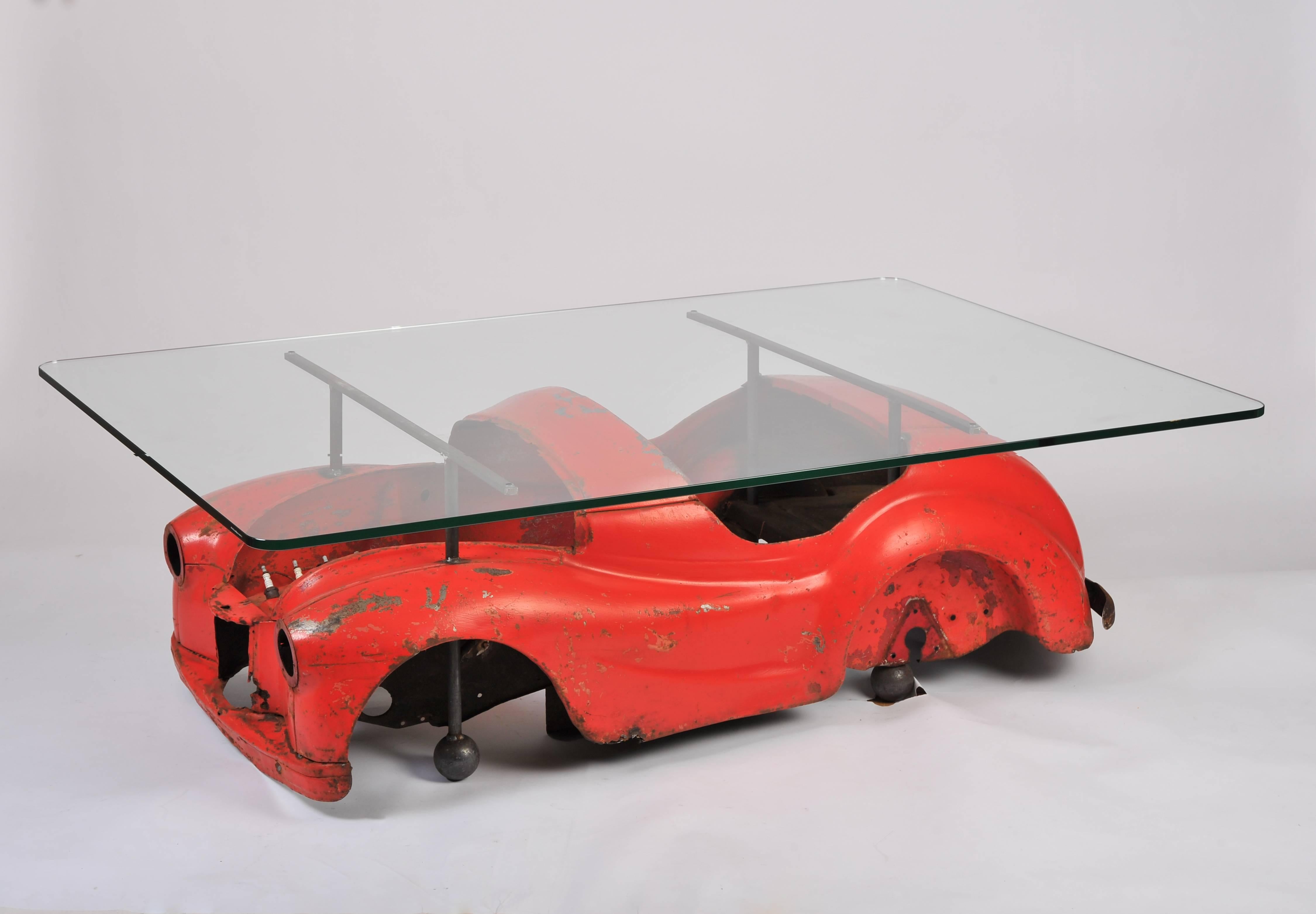 This fantastic Industrial coffee table is designed with a distressed 1950s red toy car mounted underneath a substantial glass top. The top is rectangular in shape with rounded corners. It measures 65 in - 165 cm wide, 43 1/4 in - 110 cm deep and 20