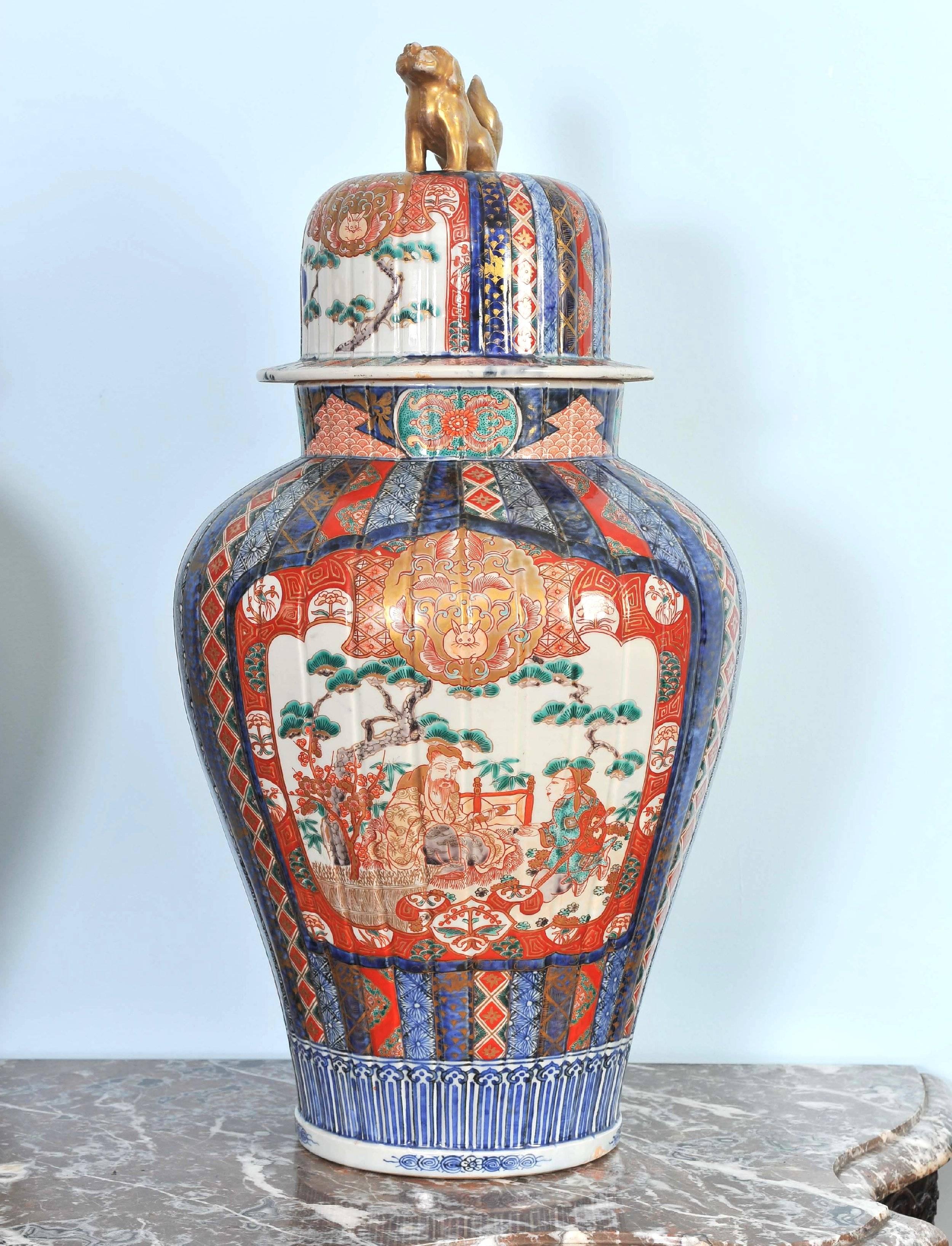 This highly decorative and fine pair of Japanese Imari ginger jars depict a central outdoor ceremonial scene surrounded by colorful geometric and floral patterned designs. The lids feature gilt painted foo dog finials and each jar has a marking on