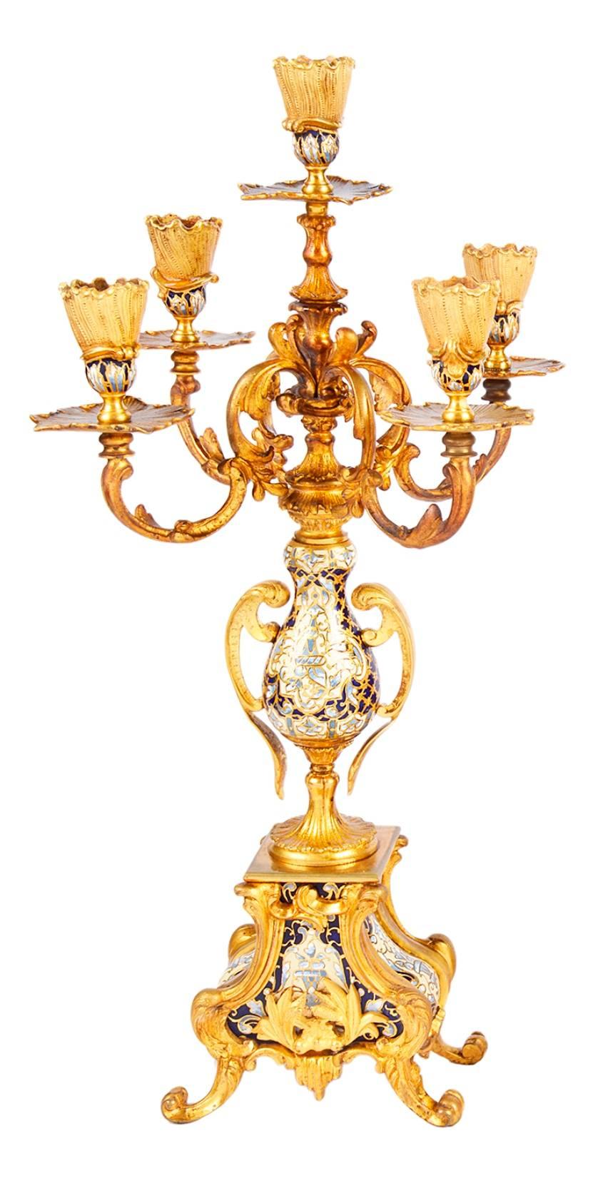 A very good quality, 19th century French gilded ormolu and champleve enamel clock garniture. The clock has a cherub on either side, an urn with swags to the top, 'C' scroll and foliate decoration and champleve enamel. The pair of candelabra each