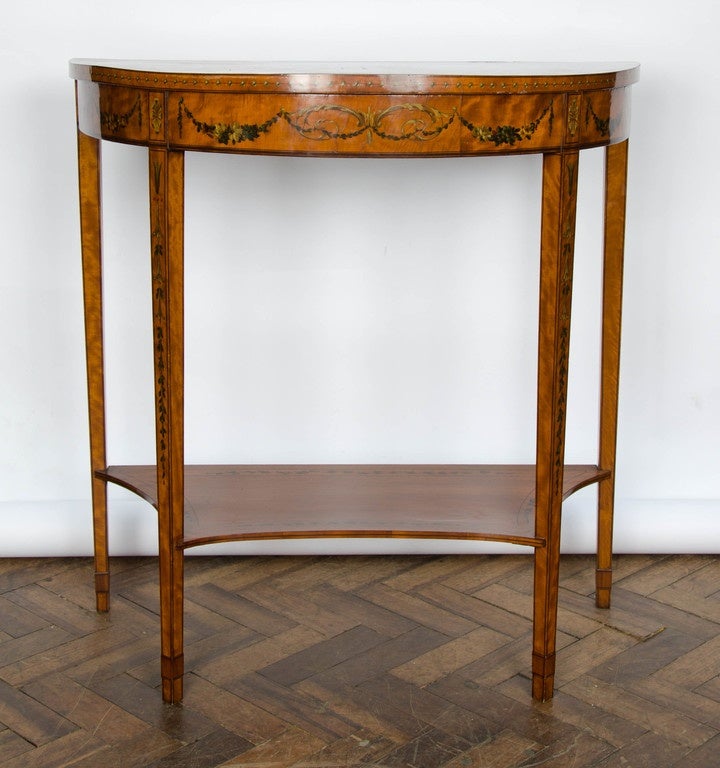 A good quality pair of Sheraton Revival satinwood demilune console tables with classical painted decoration to the top and frieze raised on square tapering legs and united by a shelf beneath.